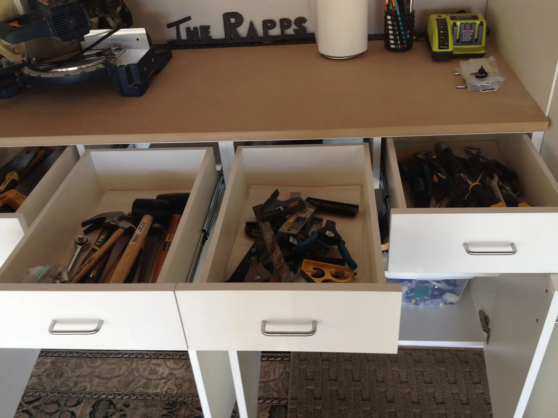 White desk with open drawers full of tools and supplies, and an open shelf above with bottles and small items