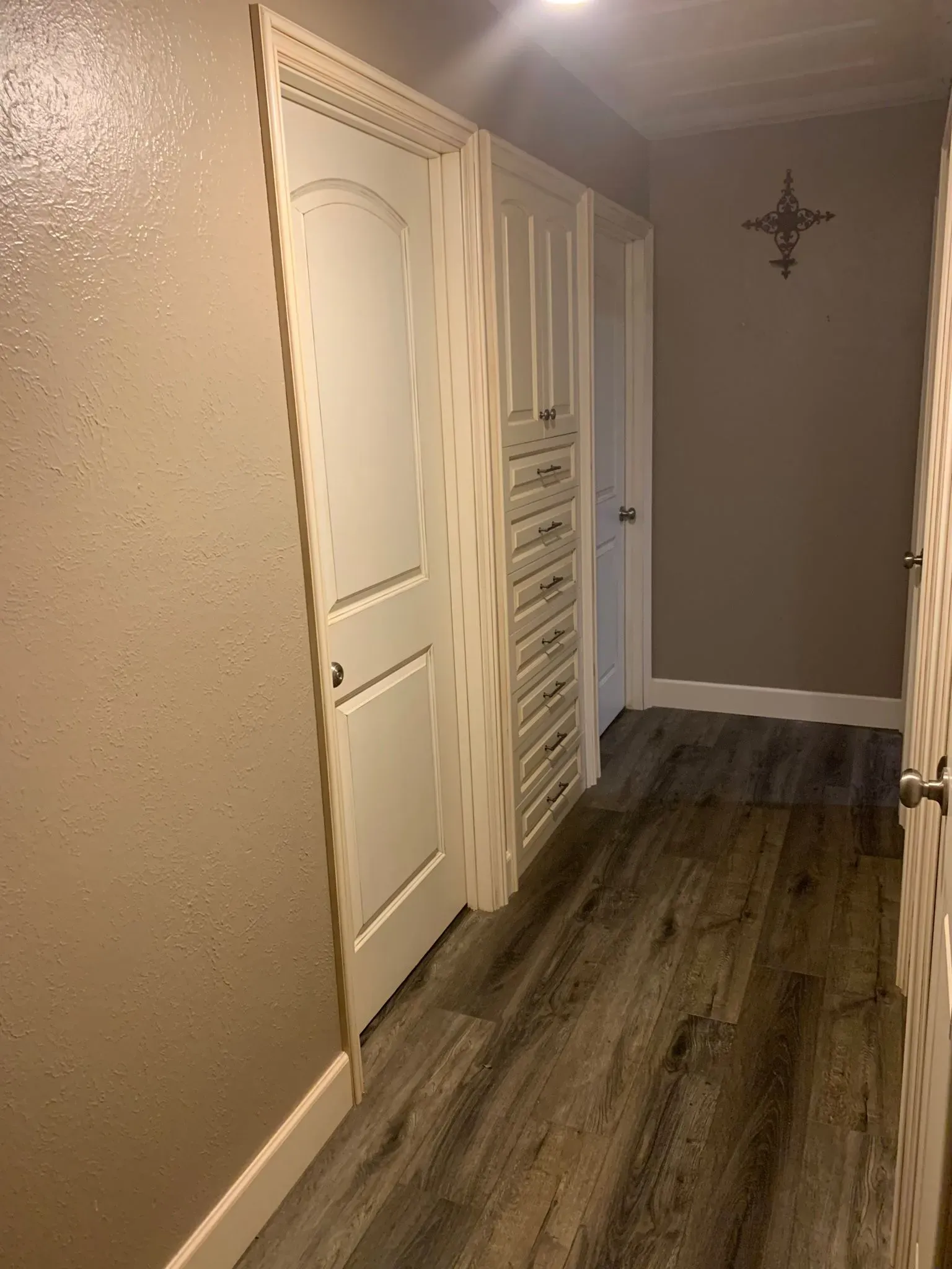 Narrow hallway with beige walls, wood-look flooring, white closet doors, and a decorative wall cross.