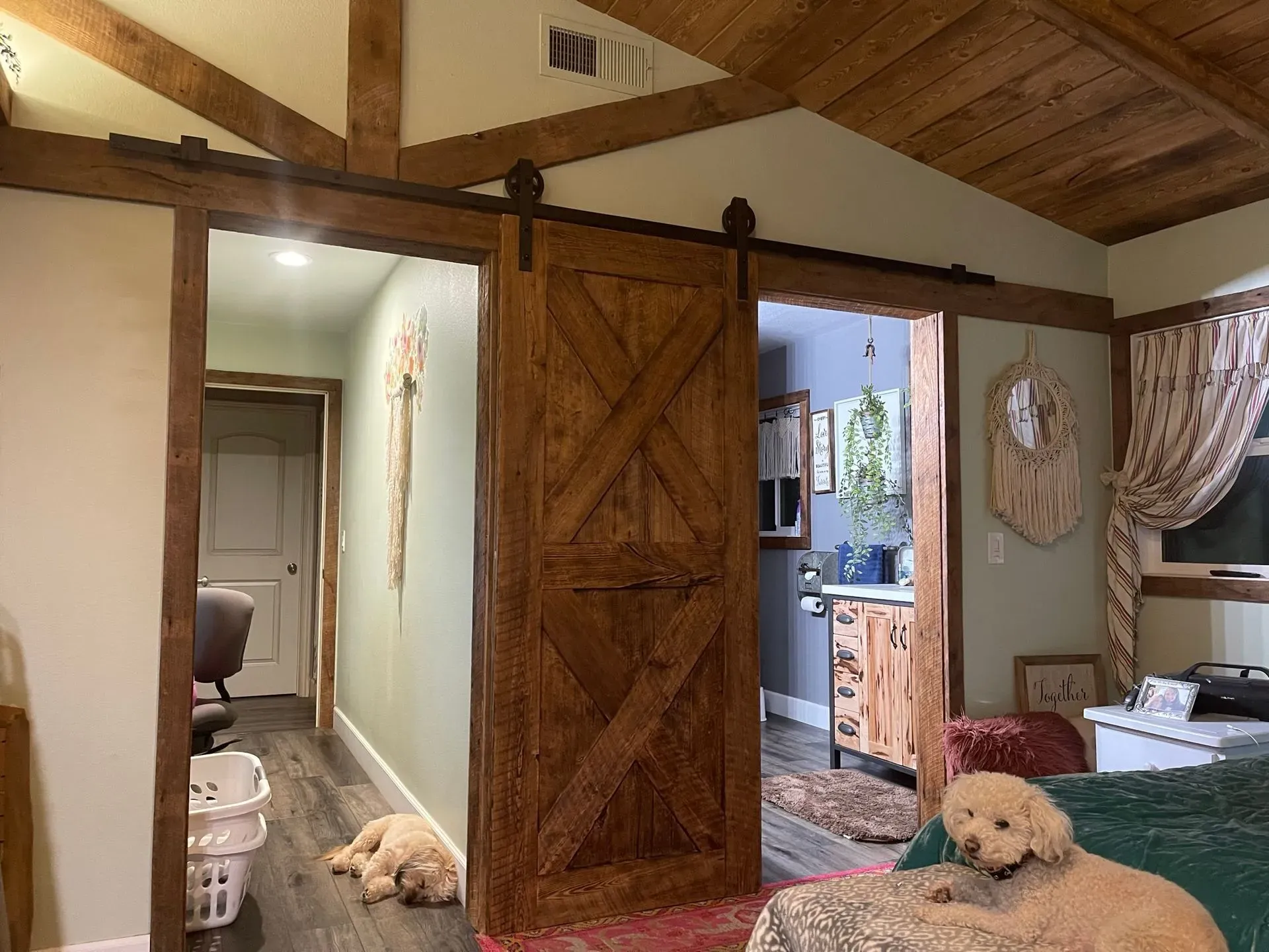 Rustic bedroom with wooden barn door, hallway, and two small white dogs lying on the floor and bed.