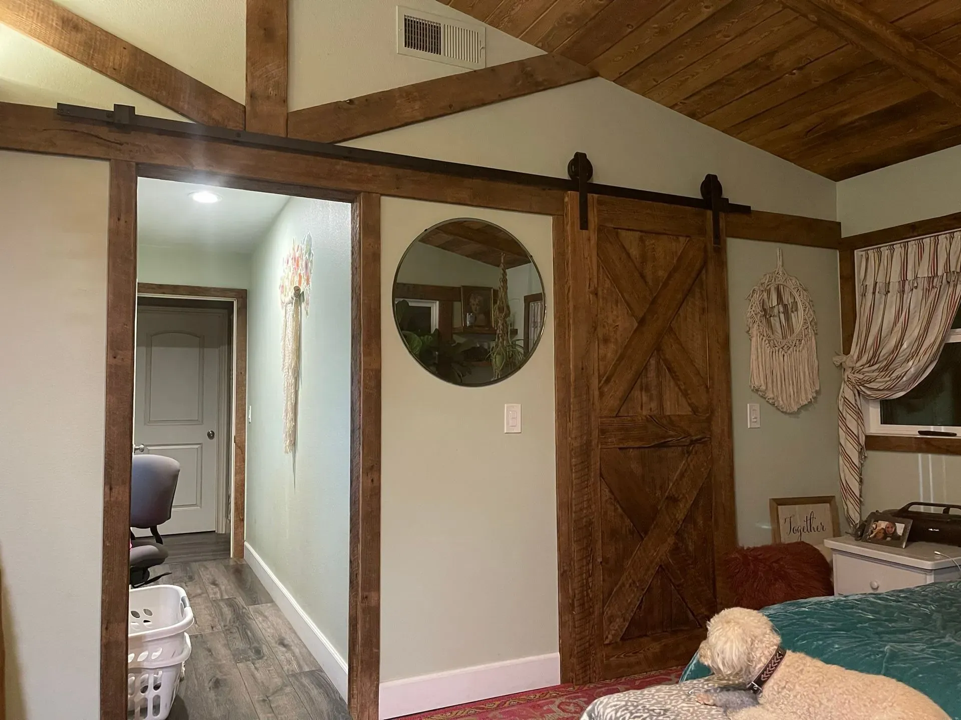 Cozy bedroom with wood-beamed ceiling, barn door, round mirror, and attached bathroom hallway