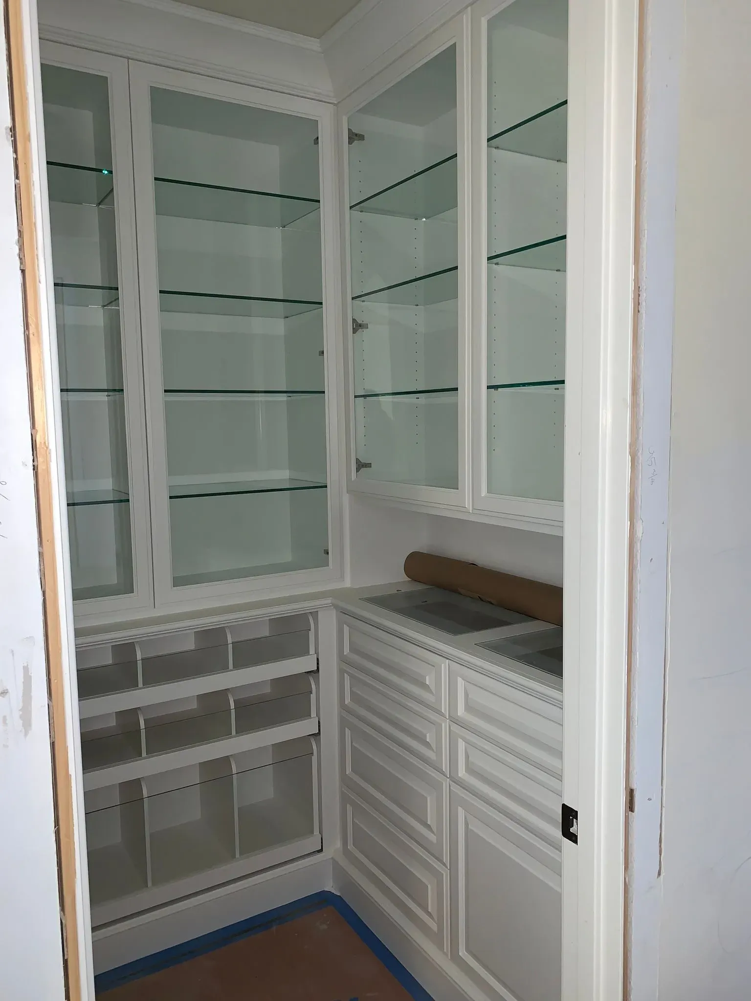 Built-in white walk-in closet with glass shelves, drawers, and a hanging rod; door open to a beige room.