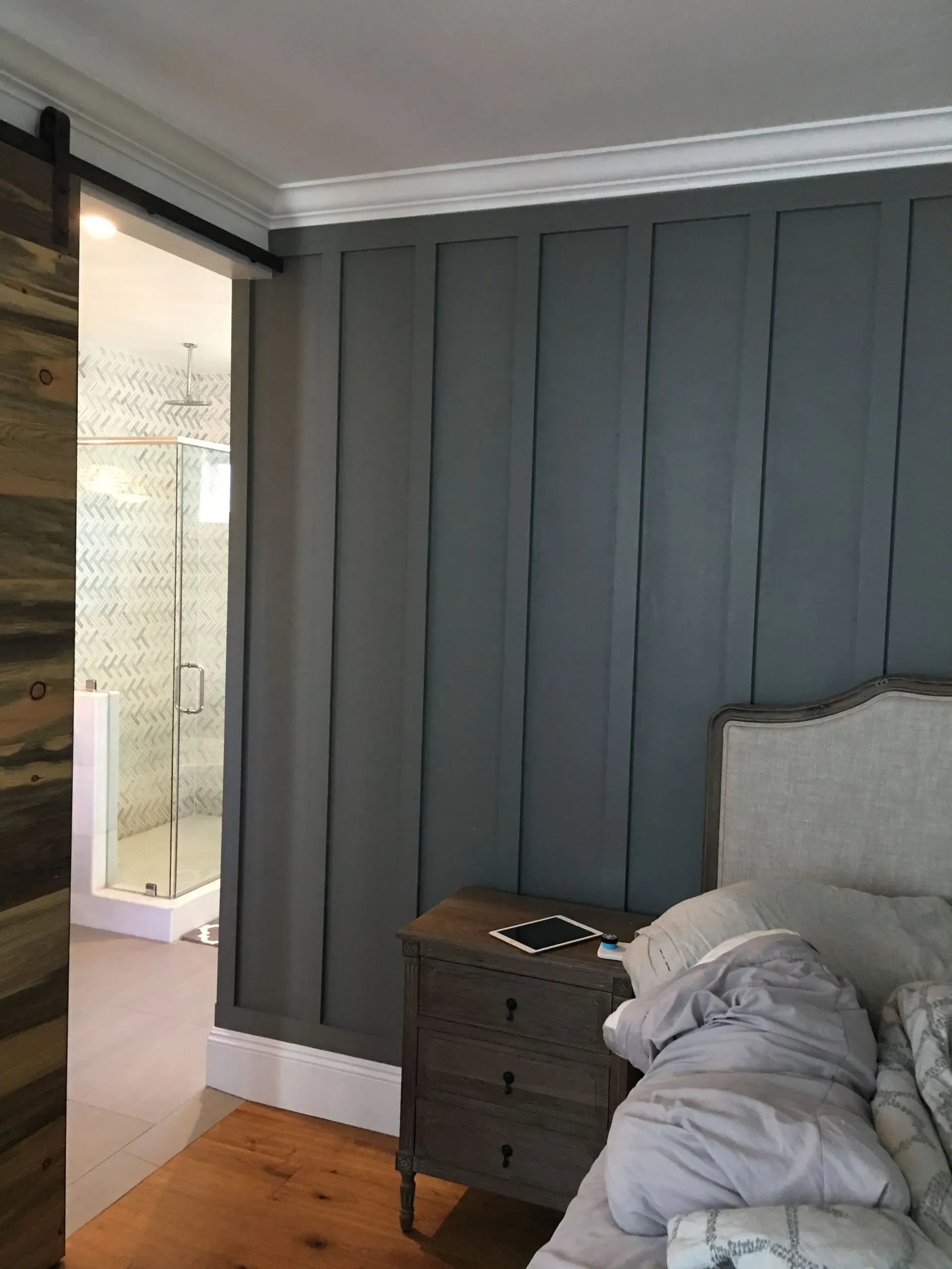 Bedroom with gray paneled wall, bed, nightstand, and open doorway to a bathroom.