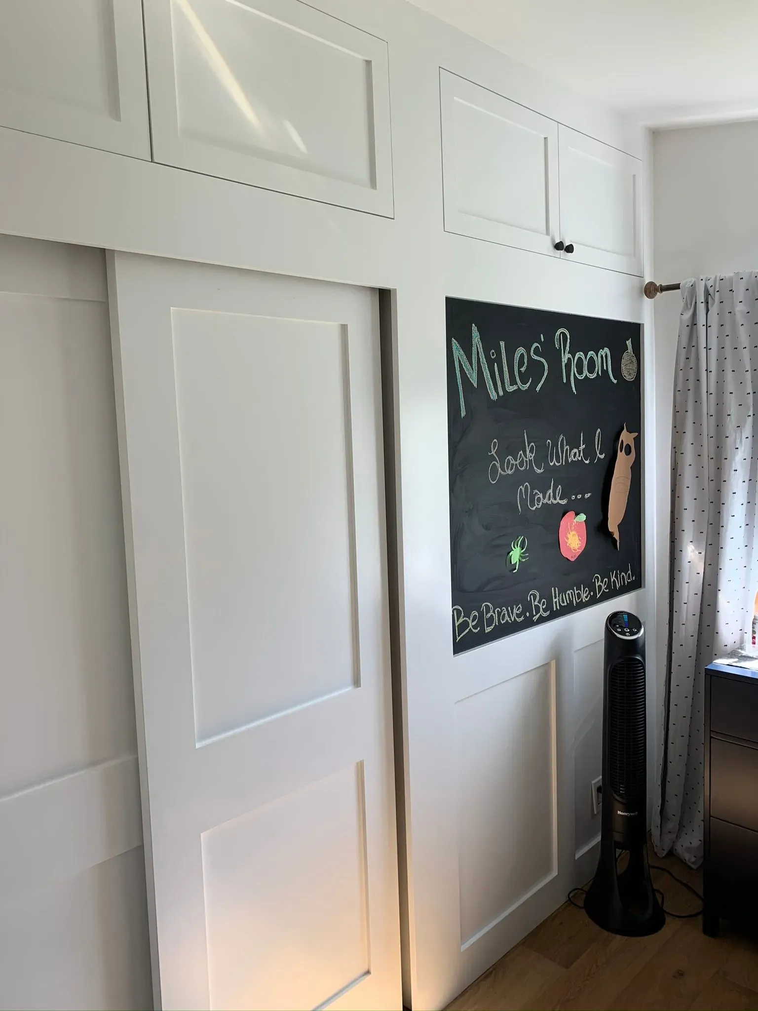 Hallway wall with a blackboard sign, white paneled doors, and a standing fan near a curtained window.