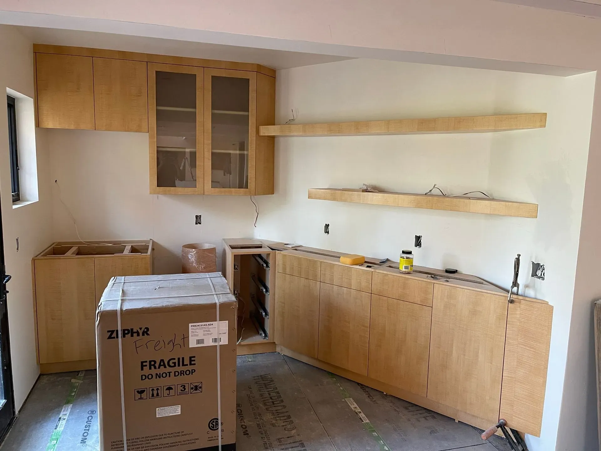 Kitchen under renovation with cabinets installed, cardboard box in foreground, and shelves mounted on bare walls.
