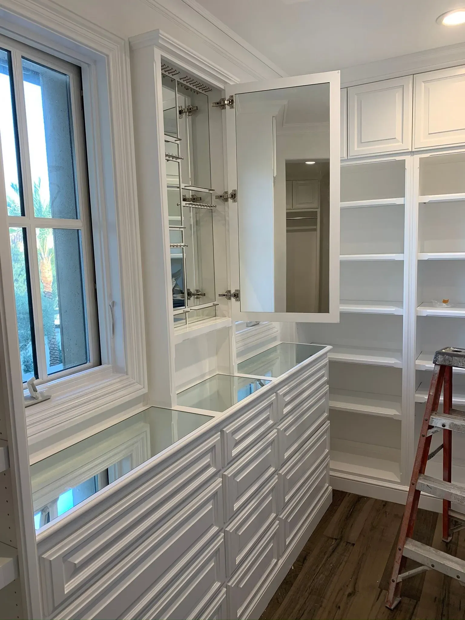 Bright white bathroom with double vanity, mirror, shelving, and a red step ladder near a window.