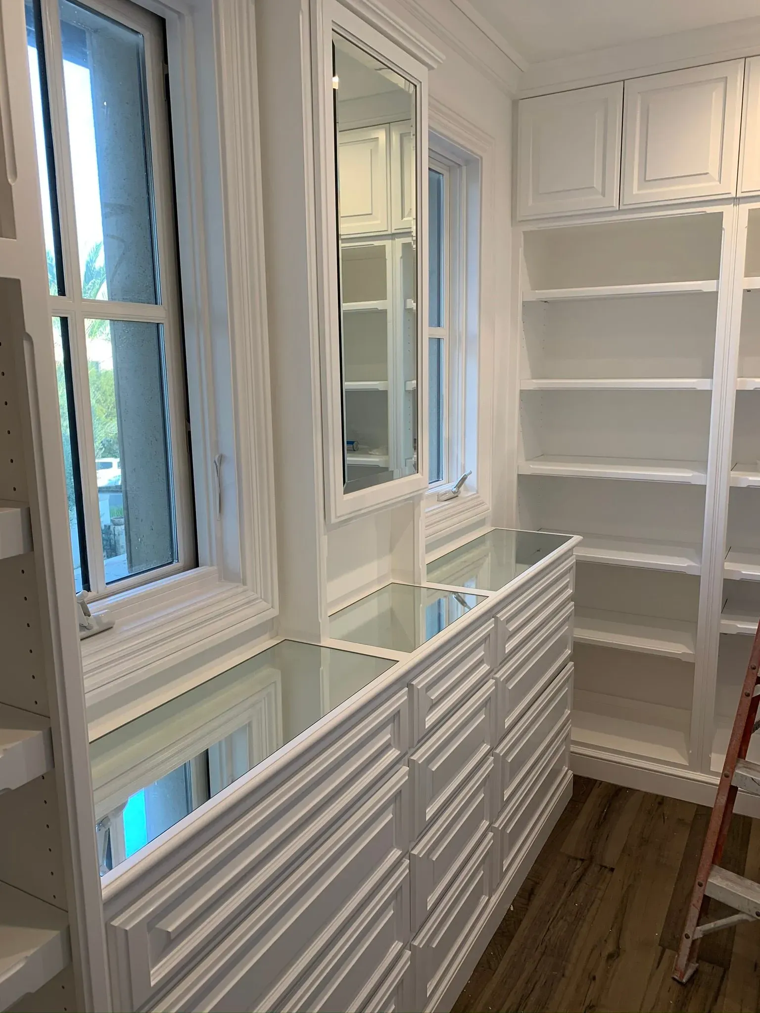 Bright white bathroom with mirrors, windows, built-in shelves, and a slatted cabinet over wood flooring