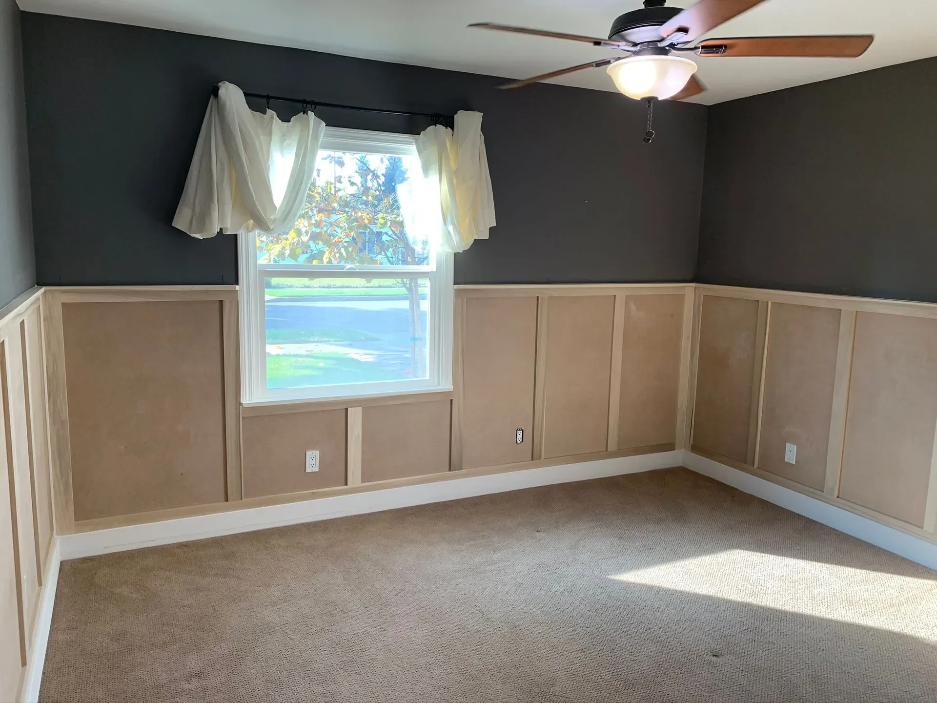 Empty bedroom with beige wainscoting, dark gray walls, a window with curtains, and carpeted floor.