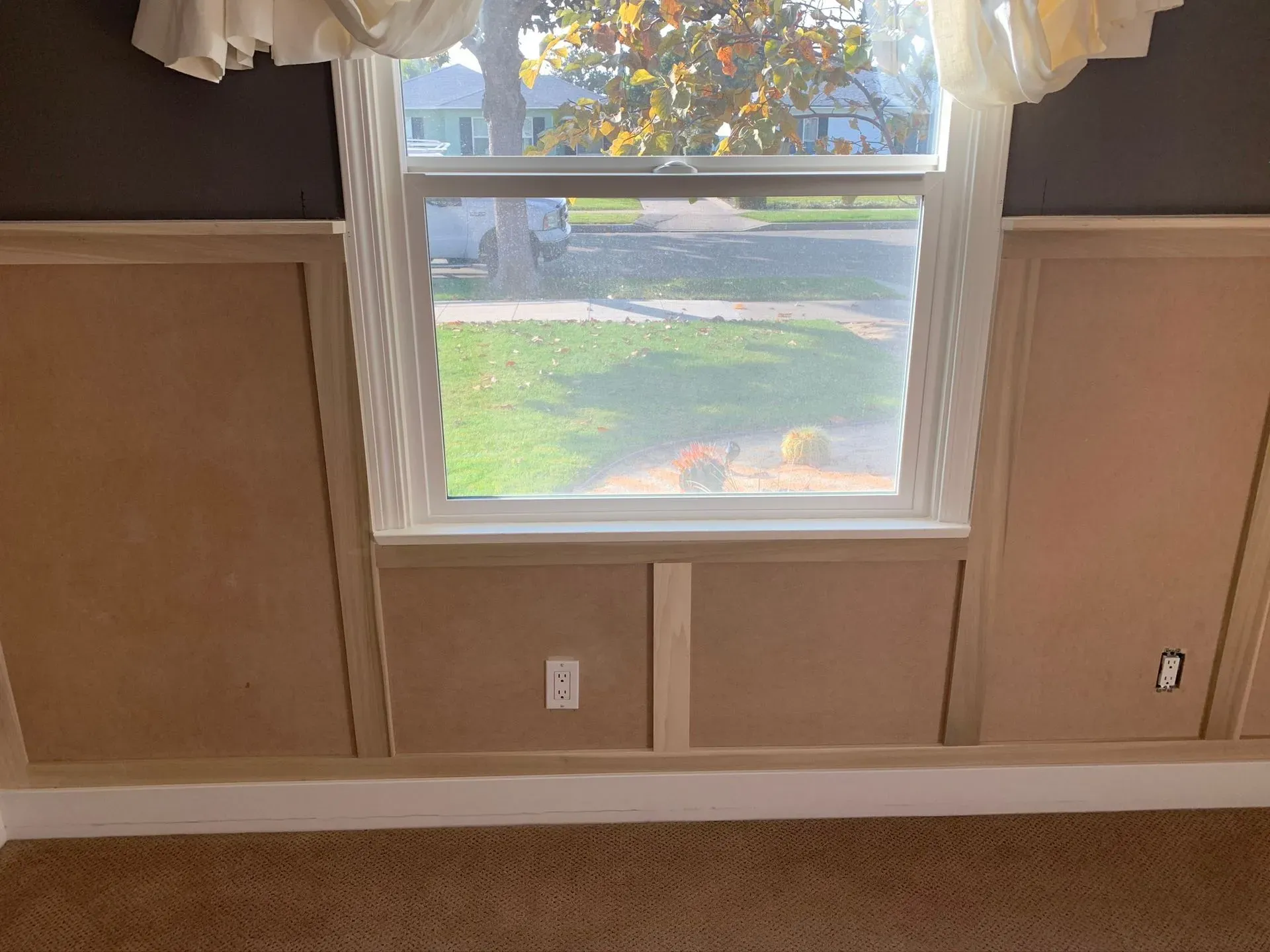 Window with white trim in a beige room, showing a yard and autumn trees outside.