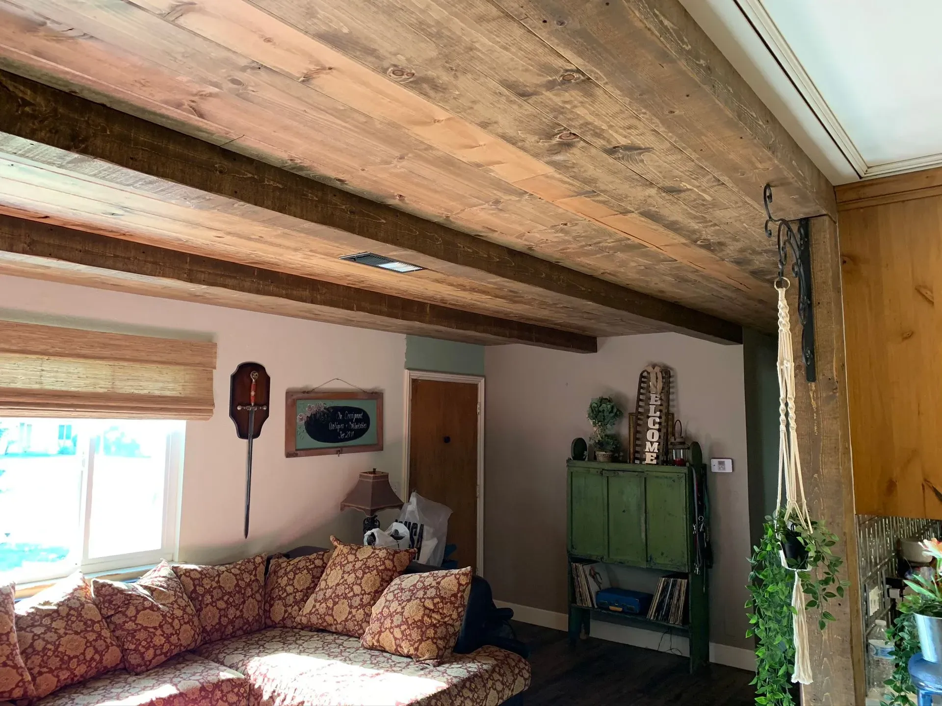 Cozy living room with exposed wooden beams, patterned sofa, window, and green cabinet against a beige wall