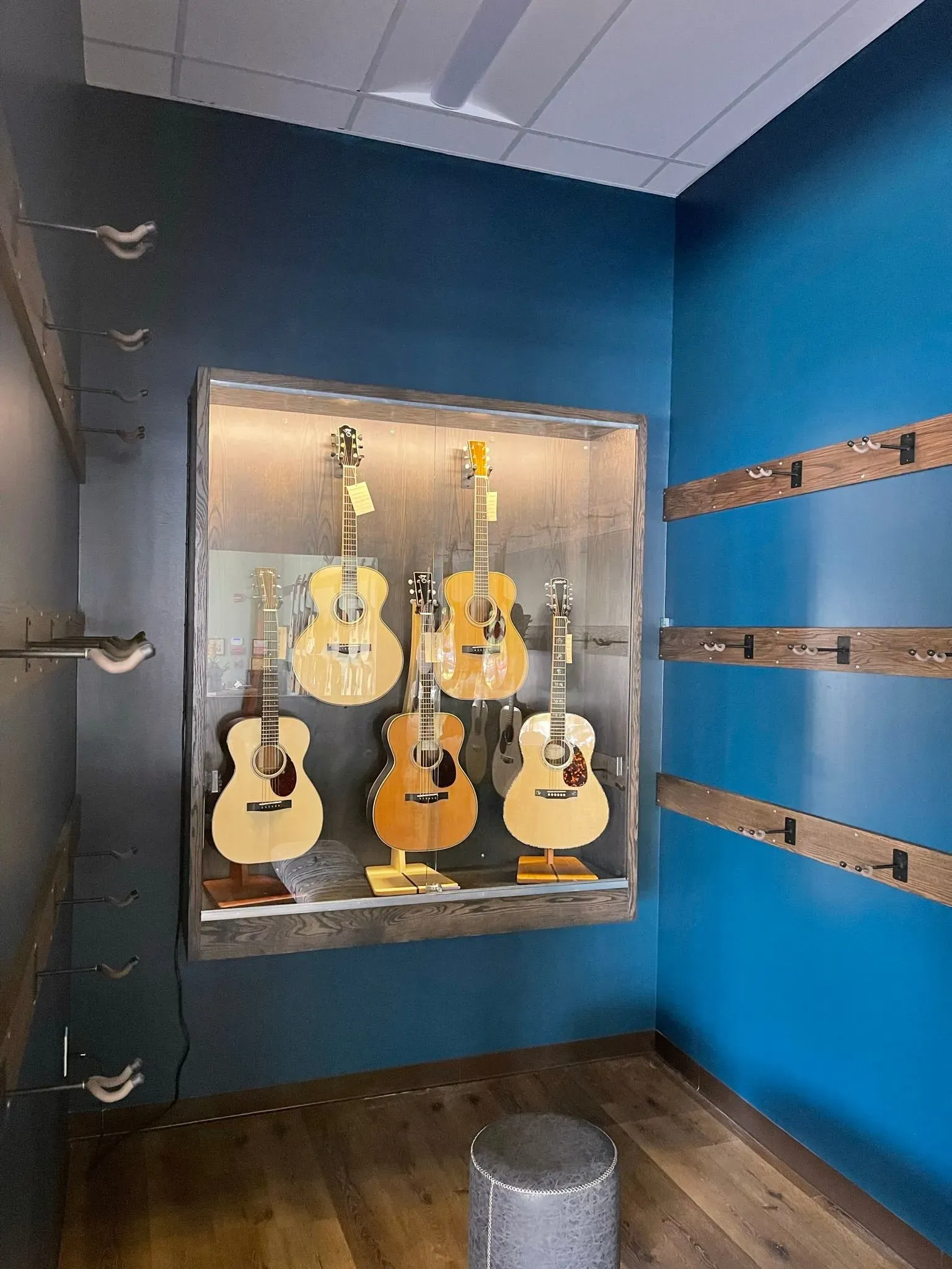 Display case of six acoustic guitars in a blue room, with wall-mounted display rails and a small stool.