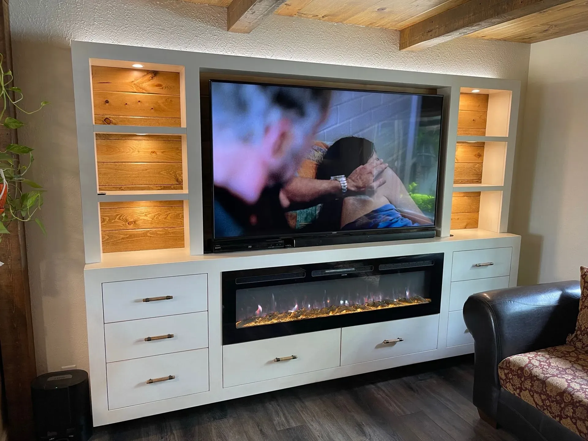 Living room media wall with built-in fireplace, TV playing a scene, and white cabinets with warm wood shelves