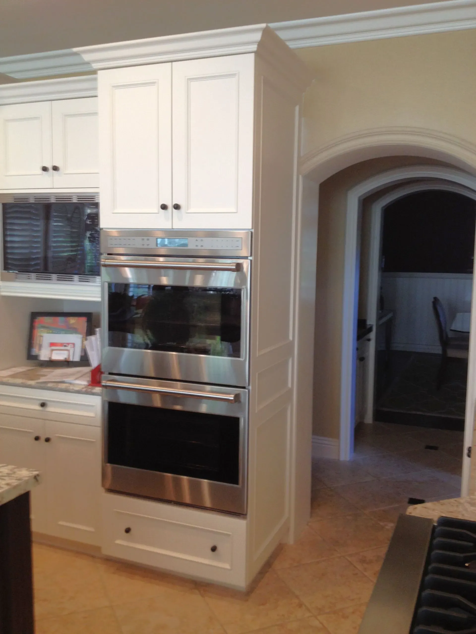 Kitchen with white cabinets, built-in stainless steel ovens, and a hallway archway in the background.