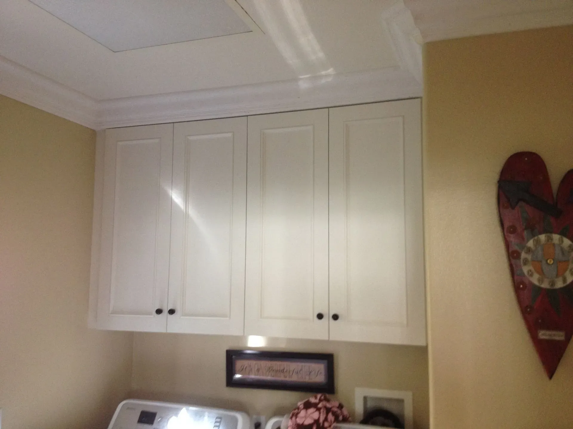 White kitchen cabinets above a countertop, with beige walls and a red heart wall decoration