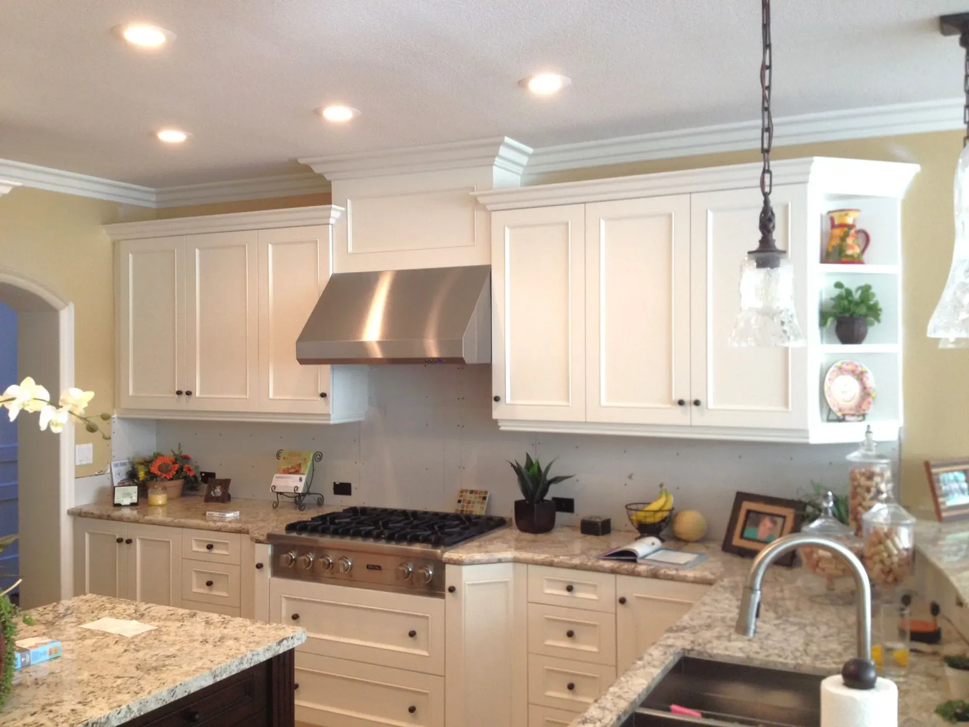 Bright white kitchen with granite island, stainless range hood, and potted plants on counters and shelves