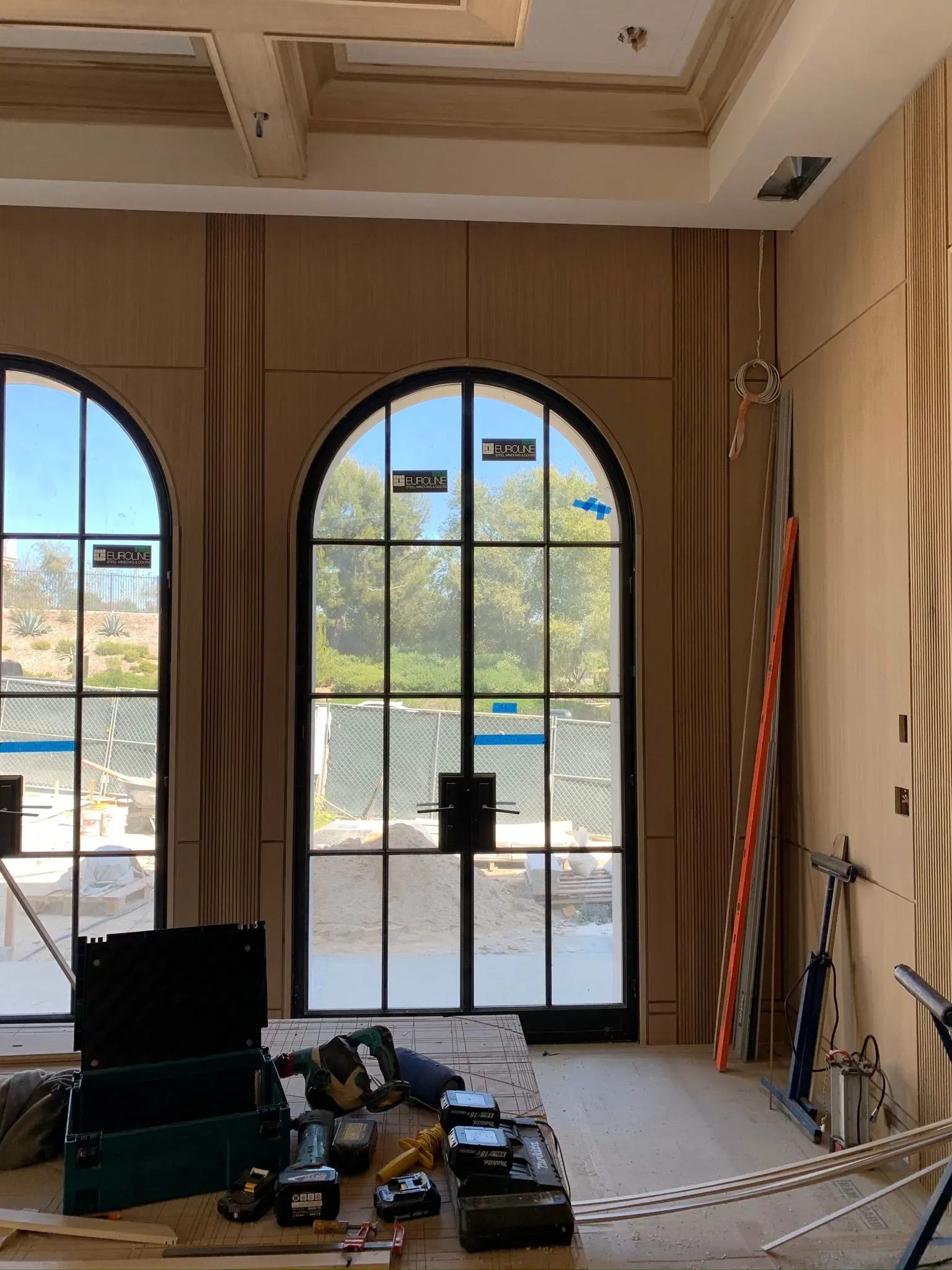 Sunlit room under renovation with arched French doors, tools on the floor, and ladders at the right side