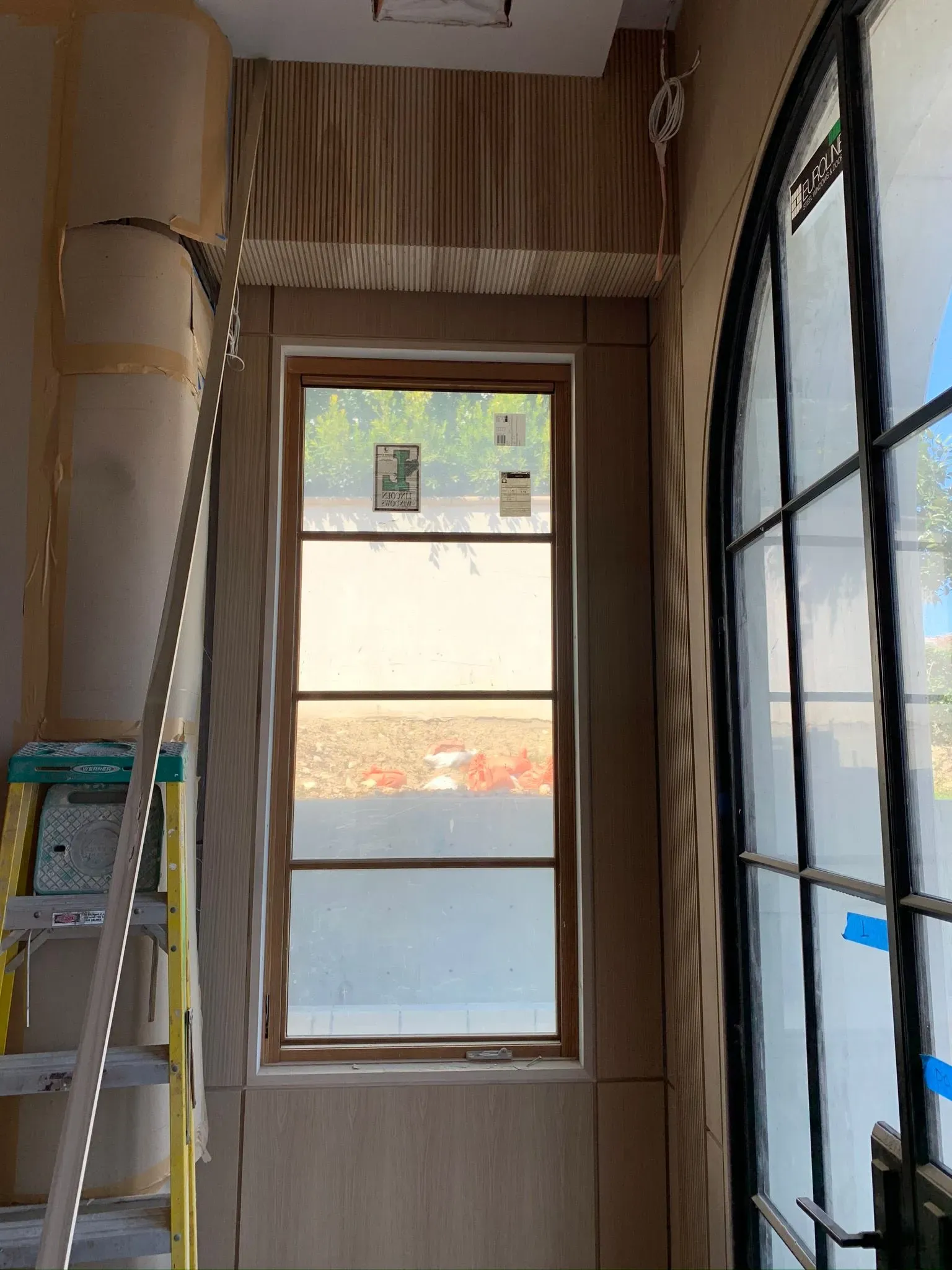 Narrow hallway with a window, ladder on the left, and a black-framed glass partition on the right.