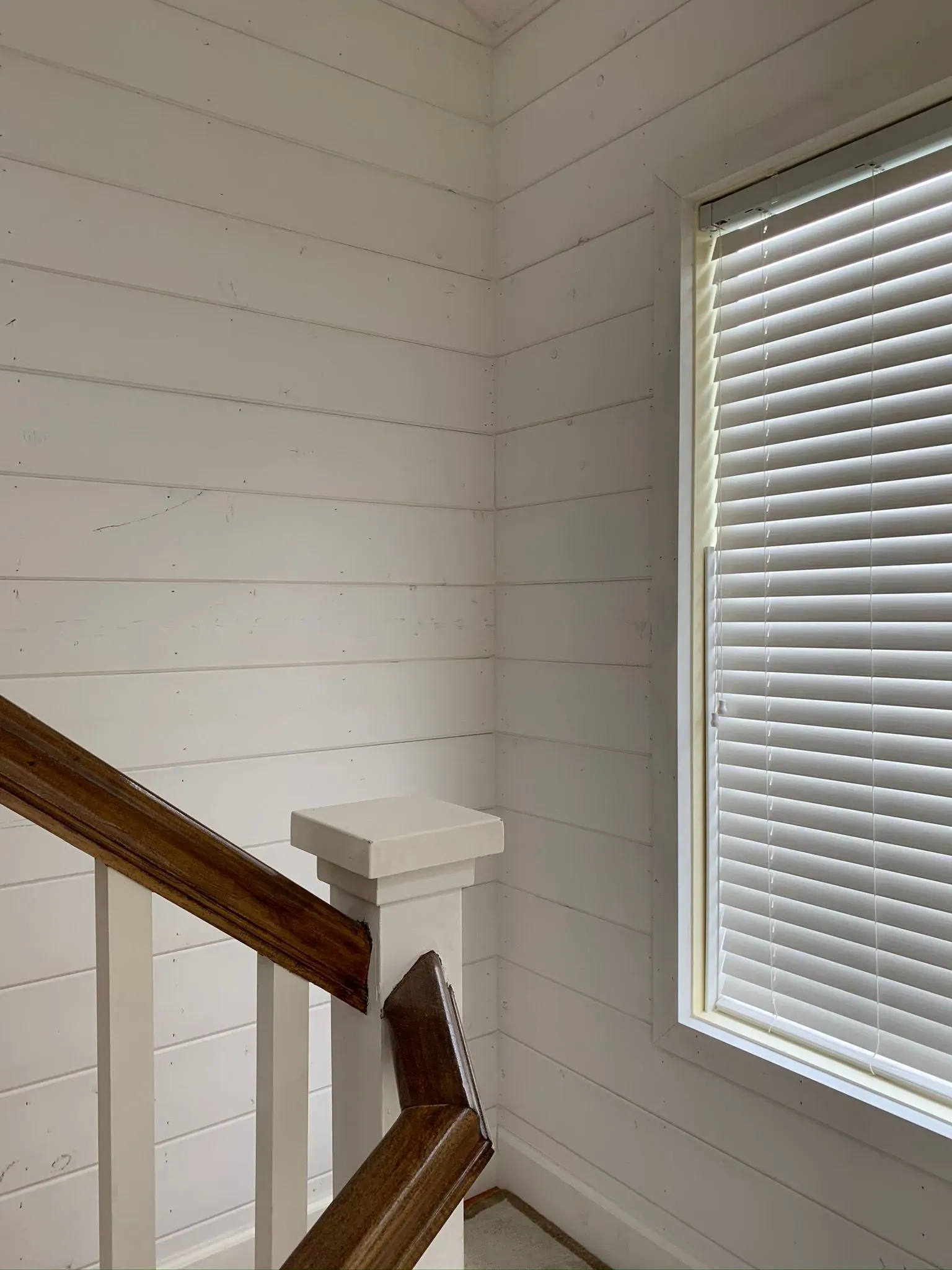 Stairwell corner with white shiplap walls, wooden handrail, and a window with closed blinds.