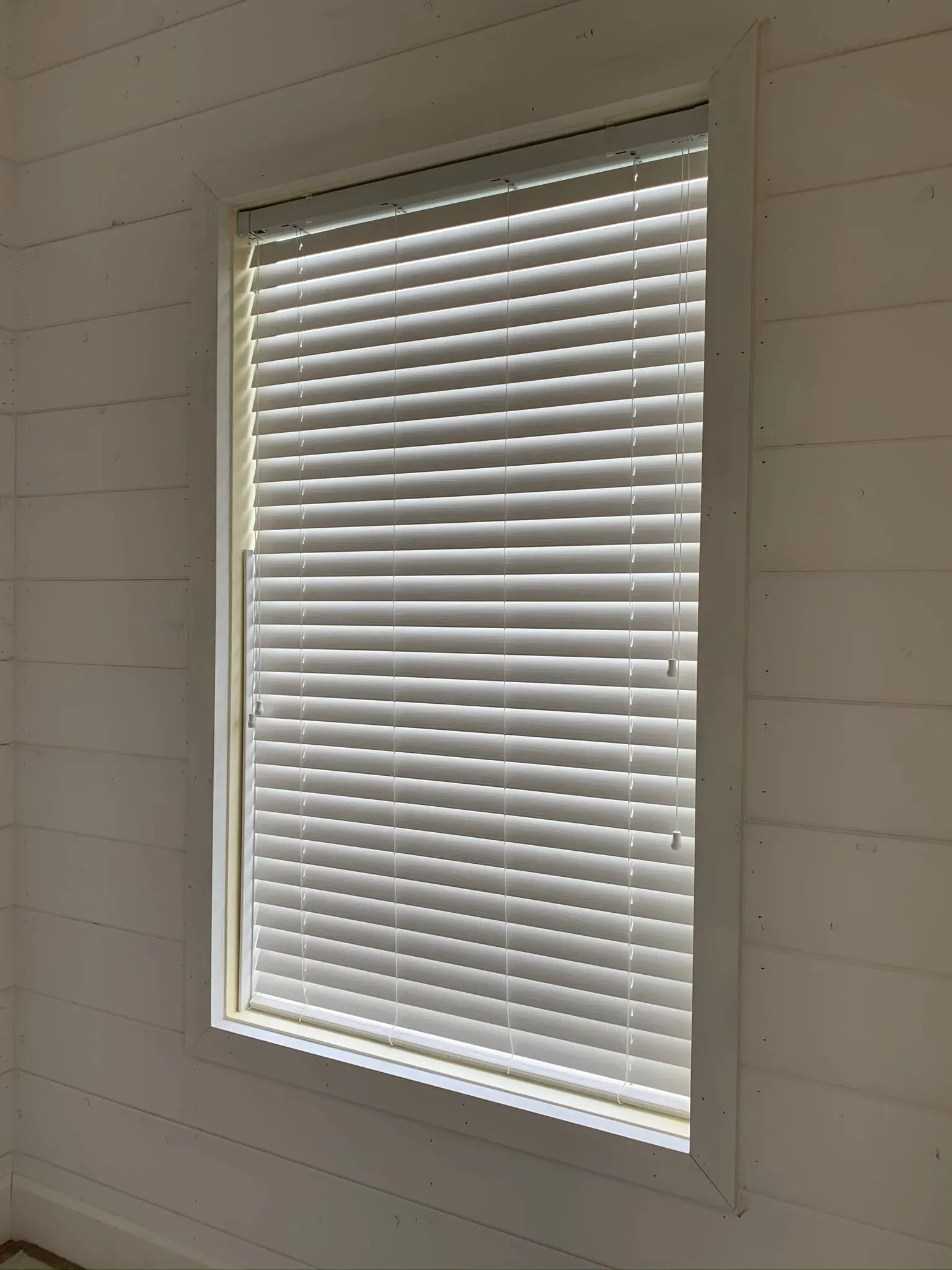 White window blinds on a small interior window set in a light paneled wall.