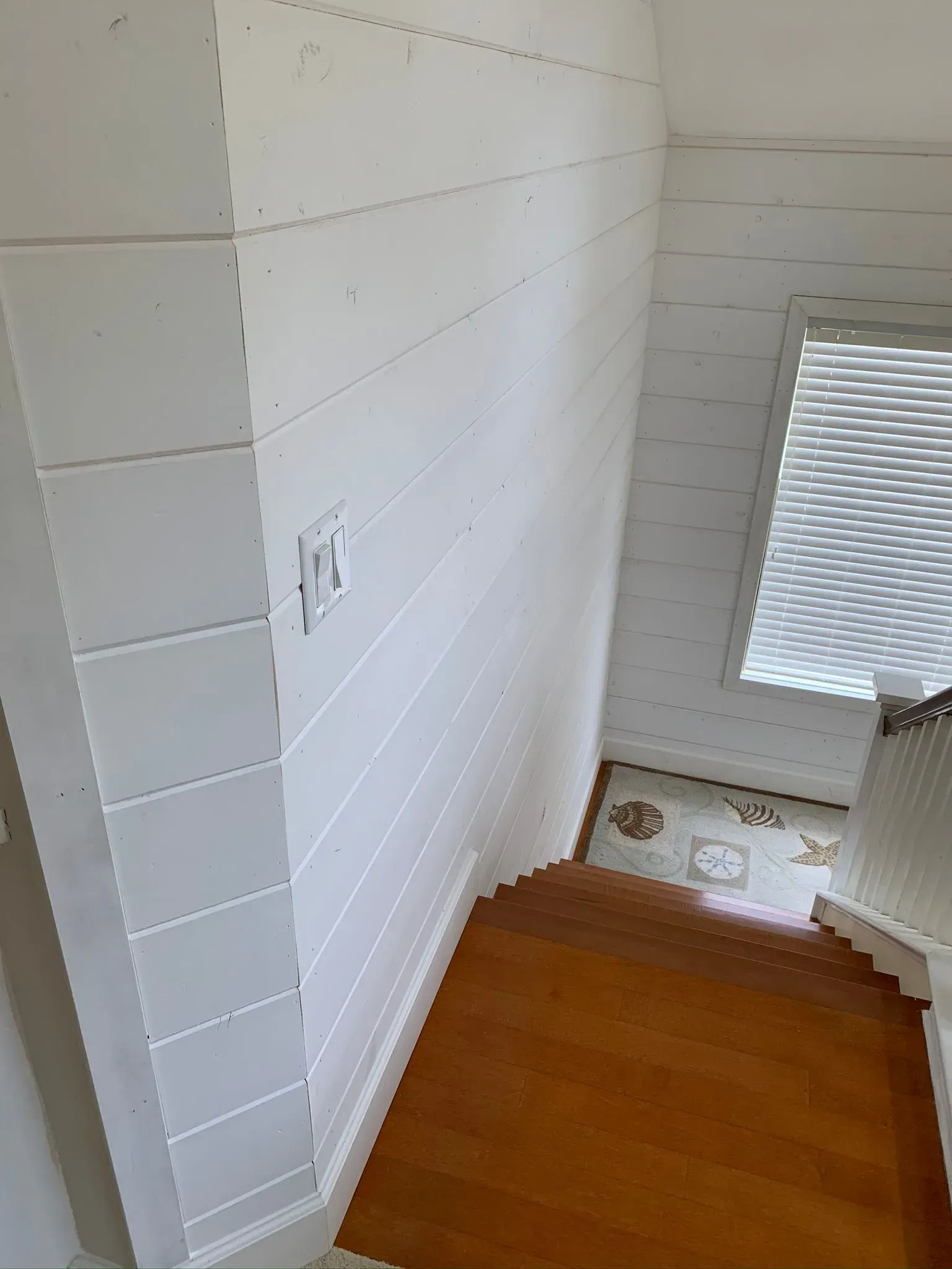 Narrow white stairwell with wooden steps and a window with blinds at the bottom