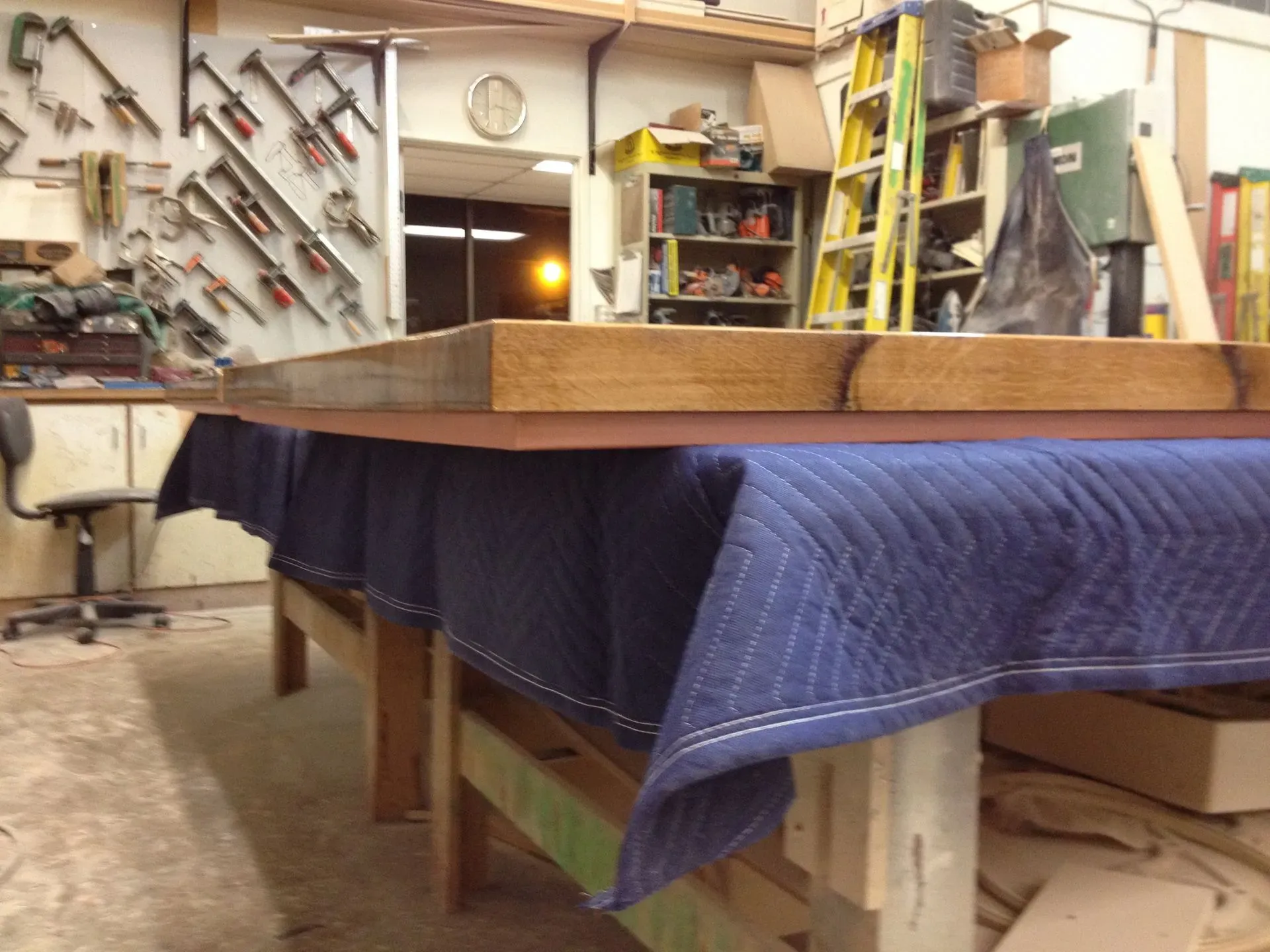 Workshop with a large wooden table covered by a blue moving blanket, tools on the wall, and shelves in the background