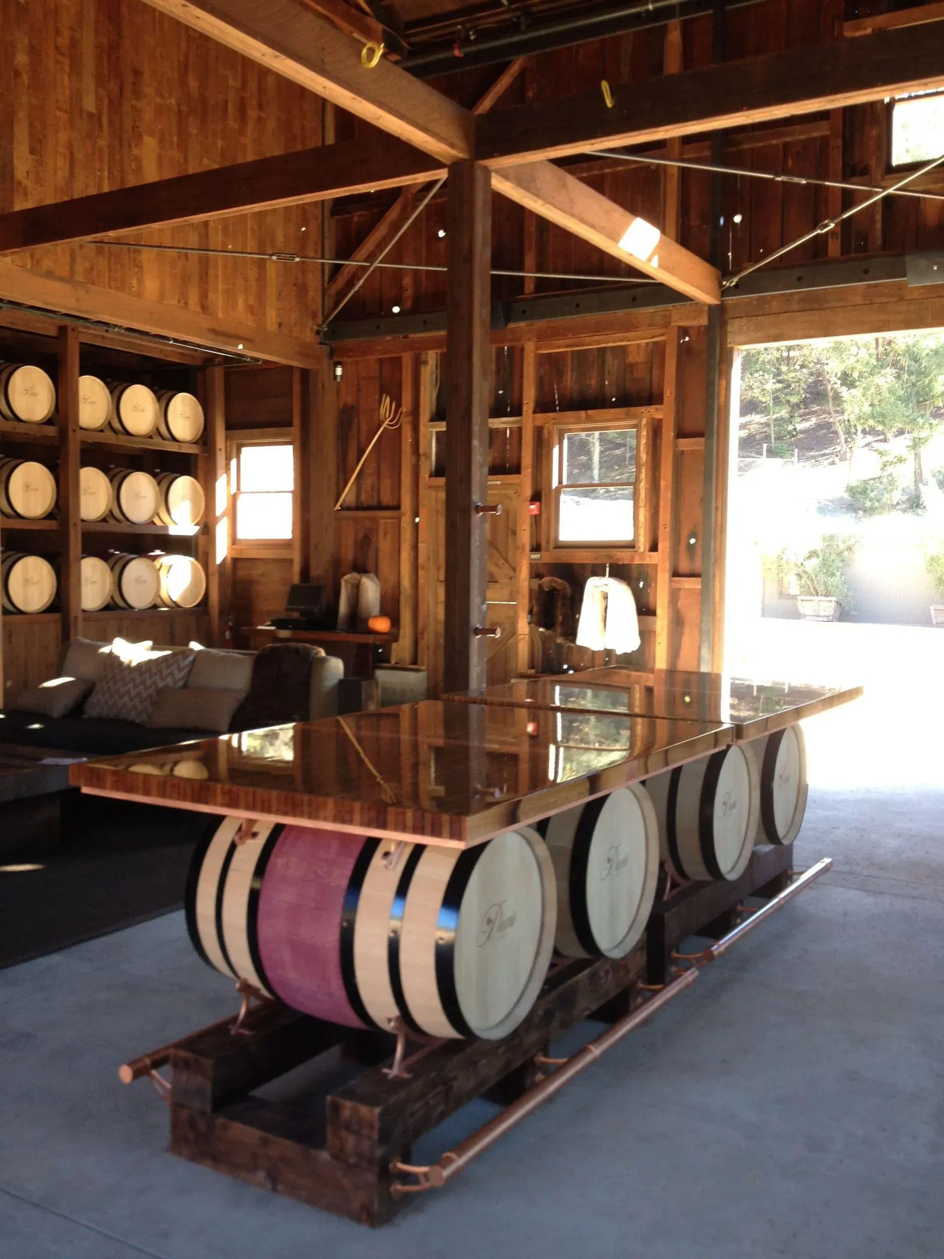 A wooden dining table made from wine barrels inside a rustic barn with barrel shelves and blue carpet.