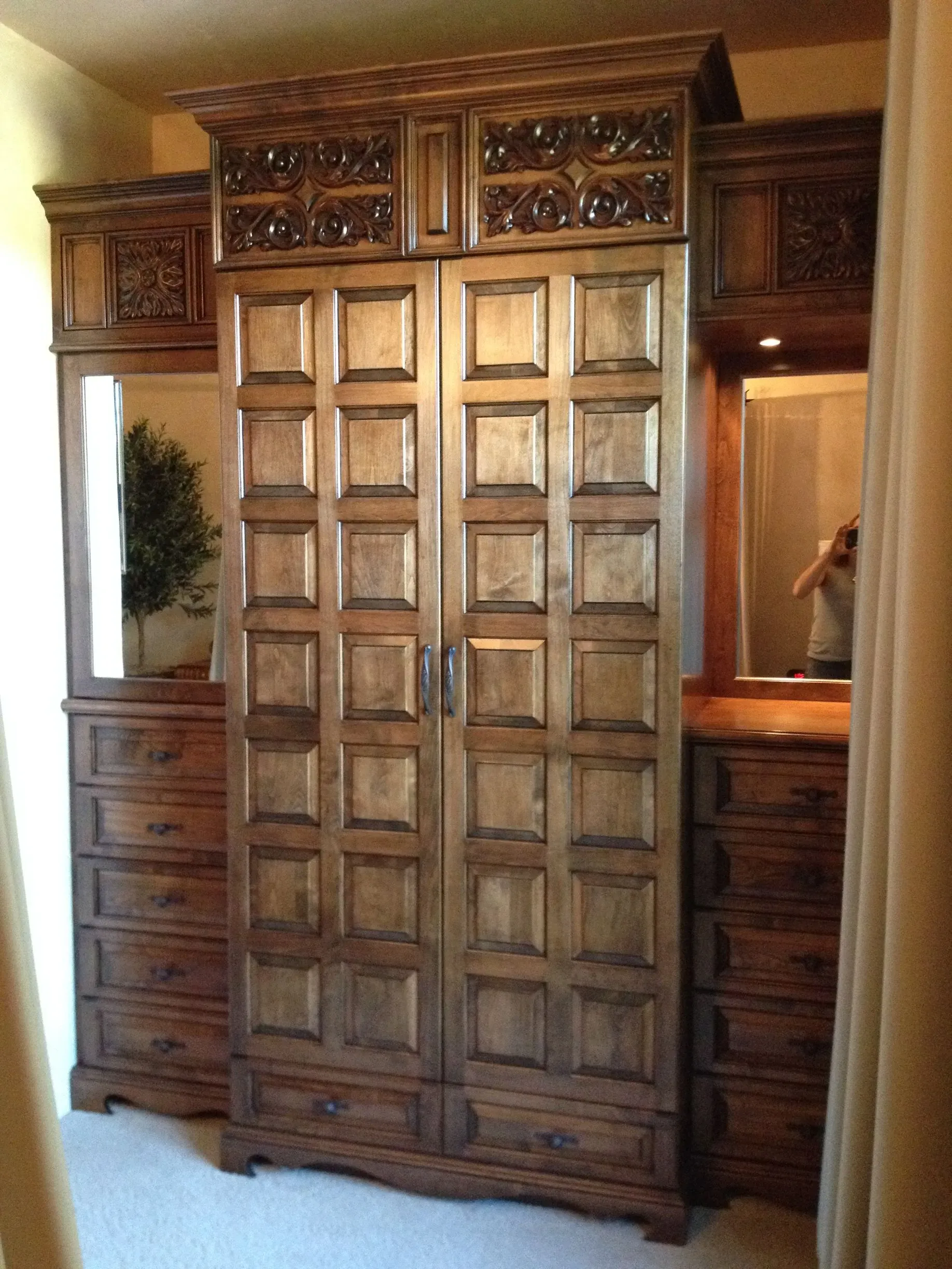 Large carved wooden armoire with paneled doors and drawers in a room, flanked by mirrors and cabinets