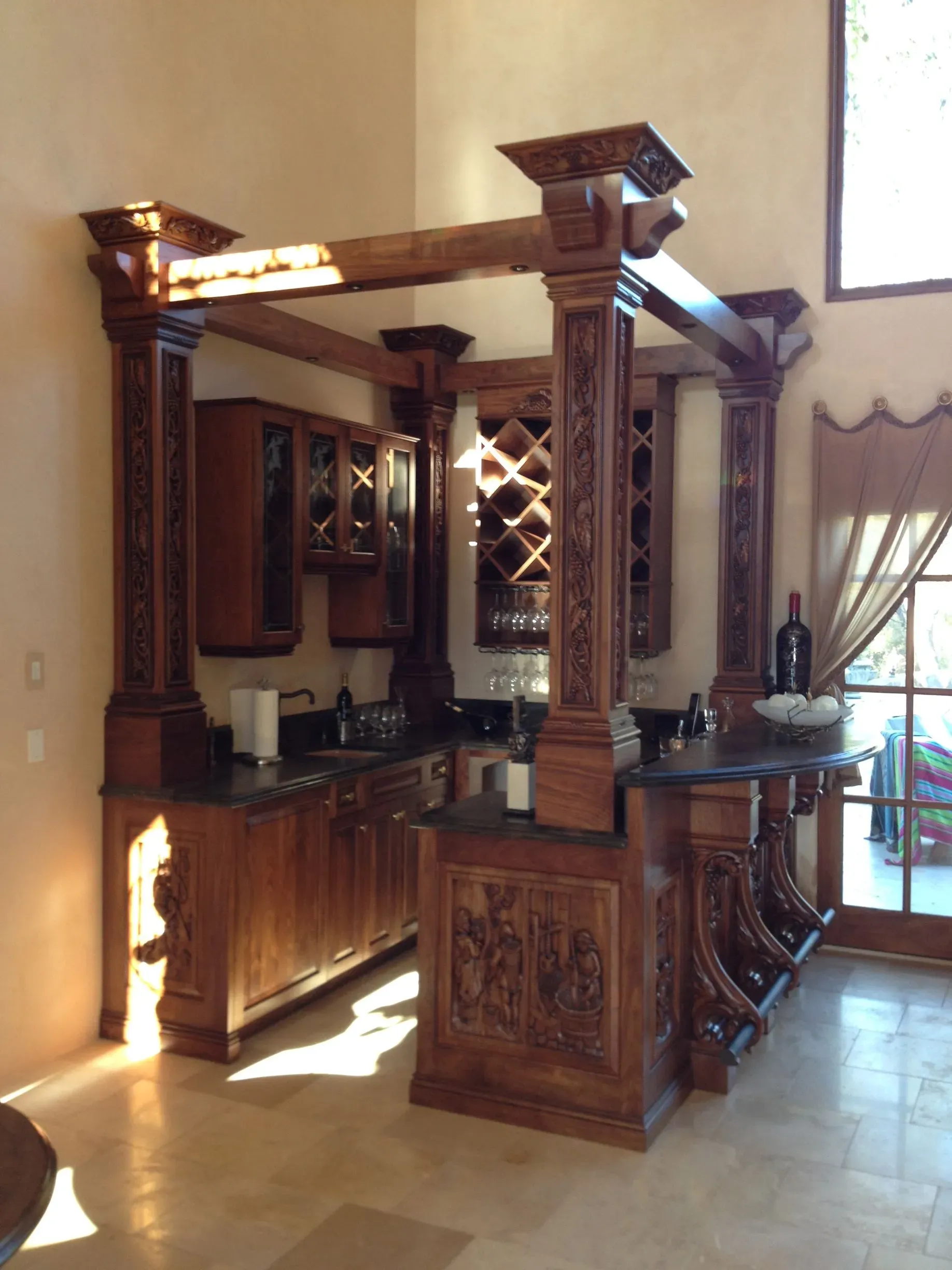 Wooden home bar with carved columns, cabinets, and a granite countertop in a sunlit room