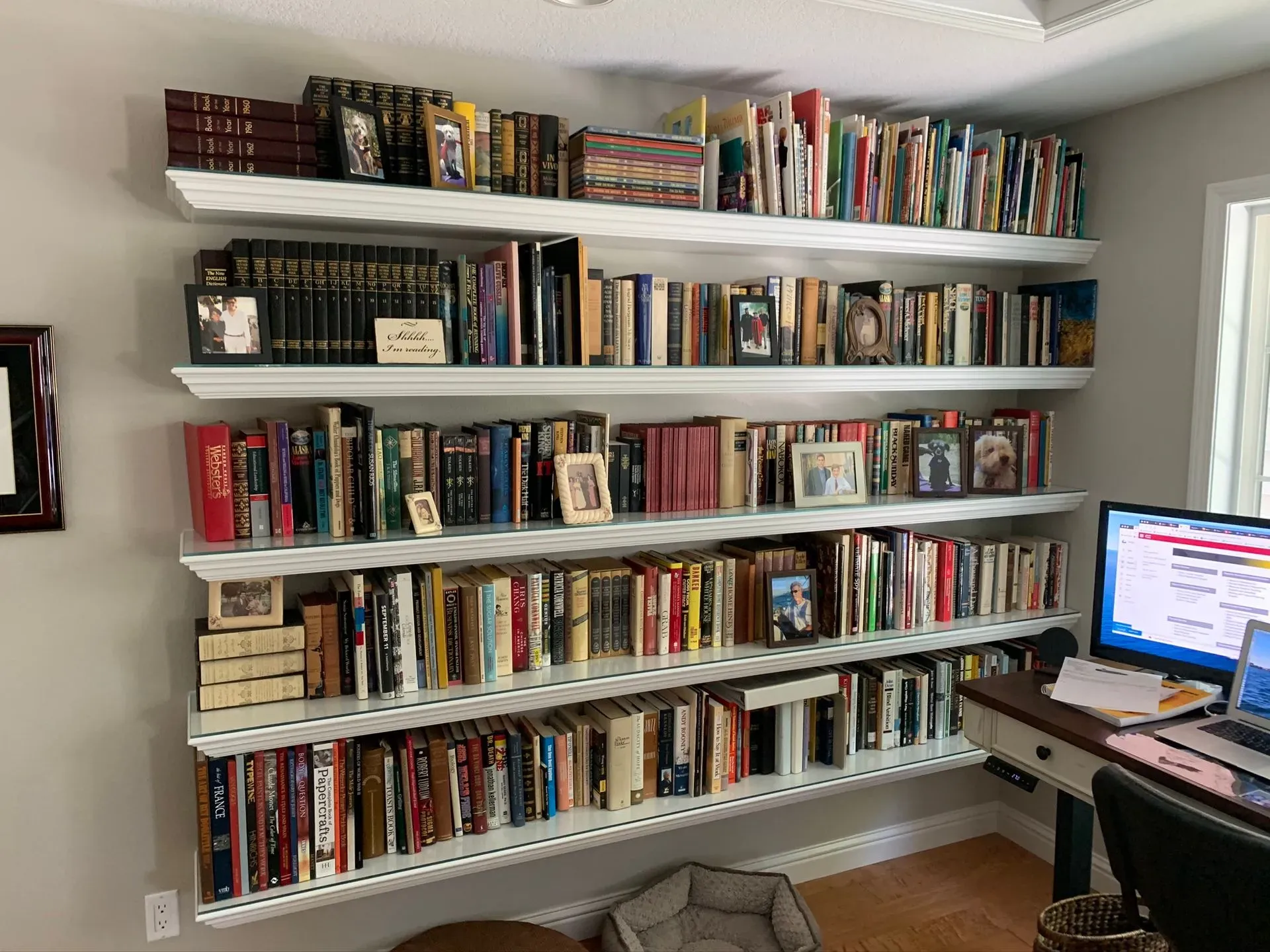 Wall of white shelves filled with books in a home office, with a desk and computer at the right.