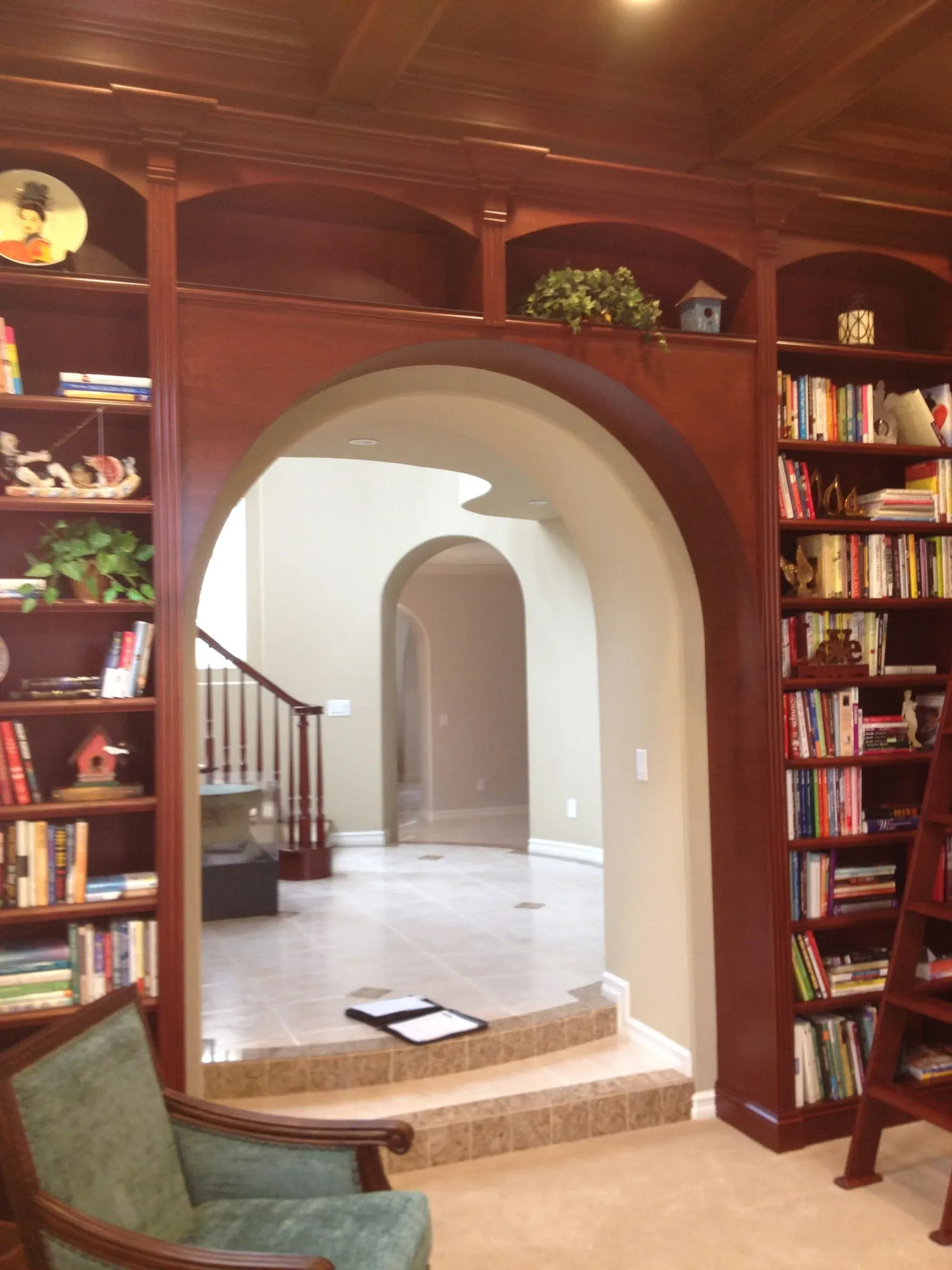 Wood-paneled library with arched doorway, bookshelves, and a green armchair overlooking a bright hallway.