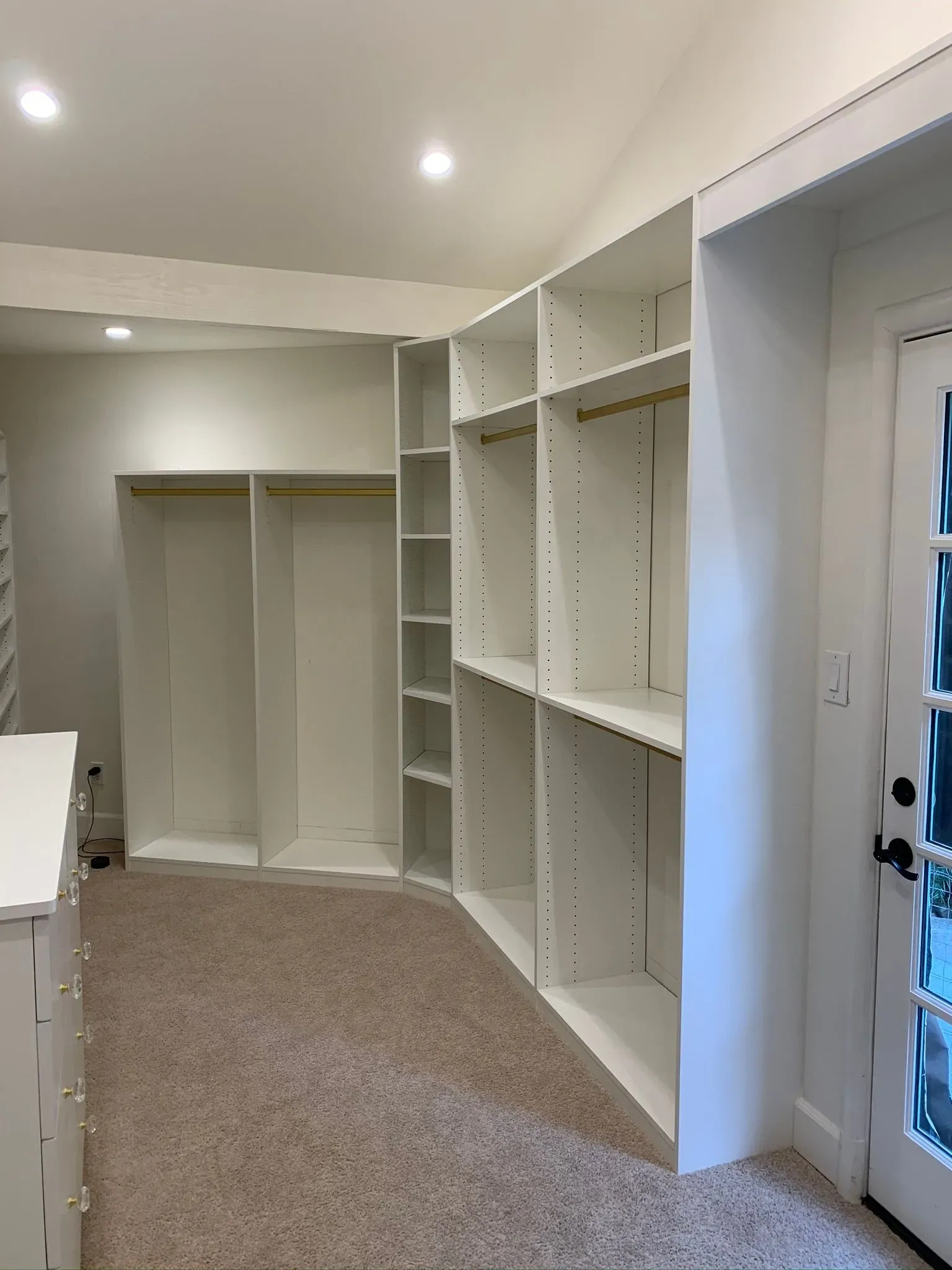 Empty walk-in closet with white built-in shelves, hanging rods, and beige carpet near a glass door