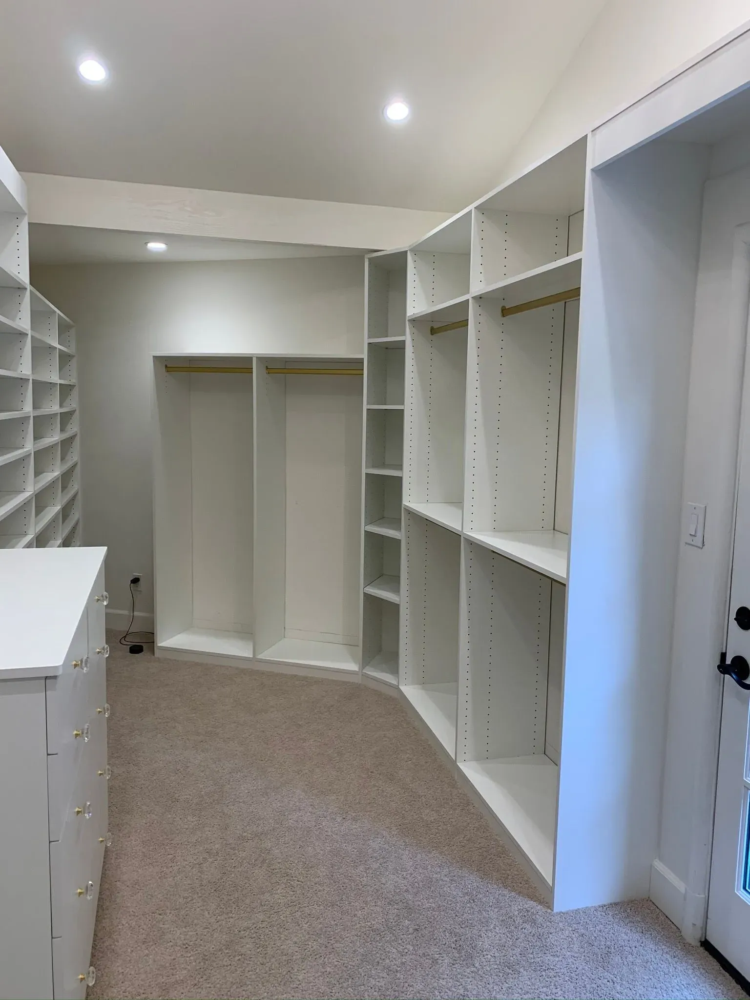 Empty walk-in closet with white built-in shelves, hanging rods, and beige carpet.