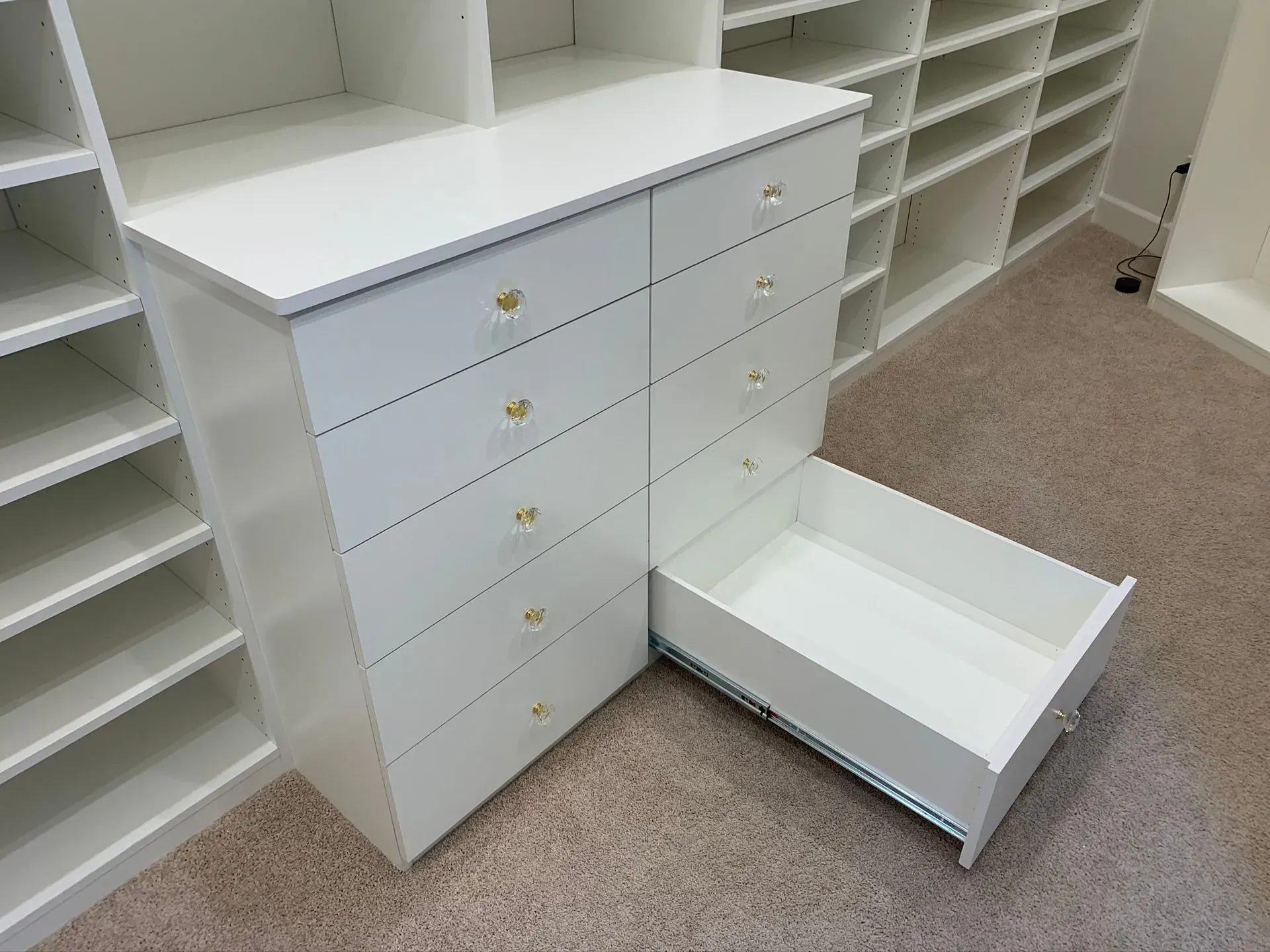 White built-in drawers and shelves in a closet, with one drawer pulled out on beige carpet.