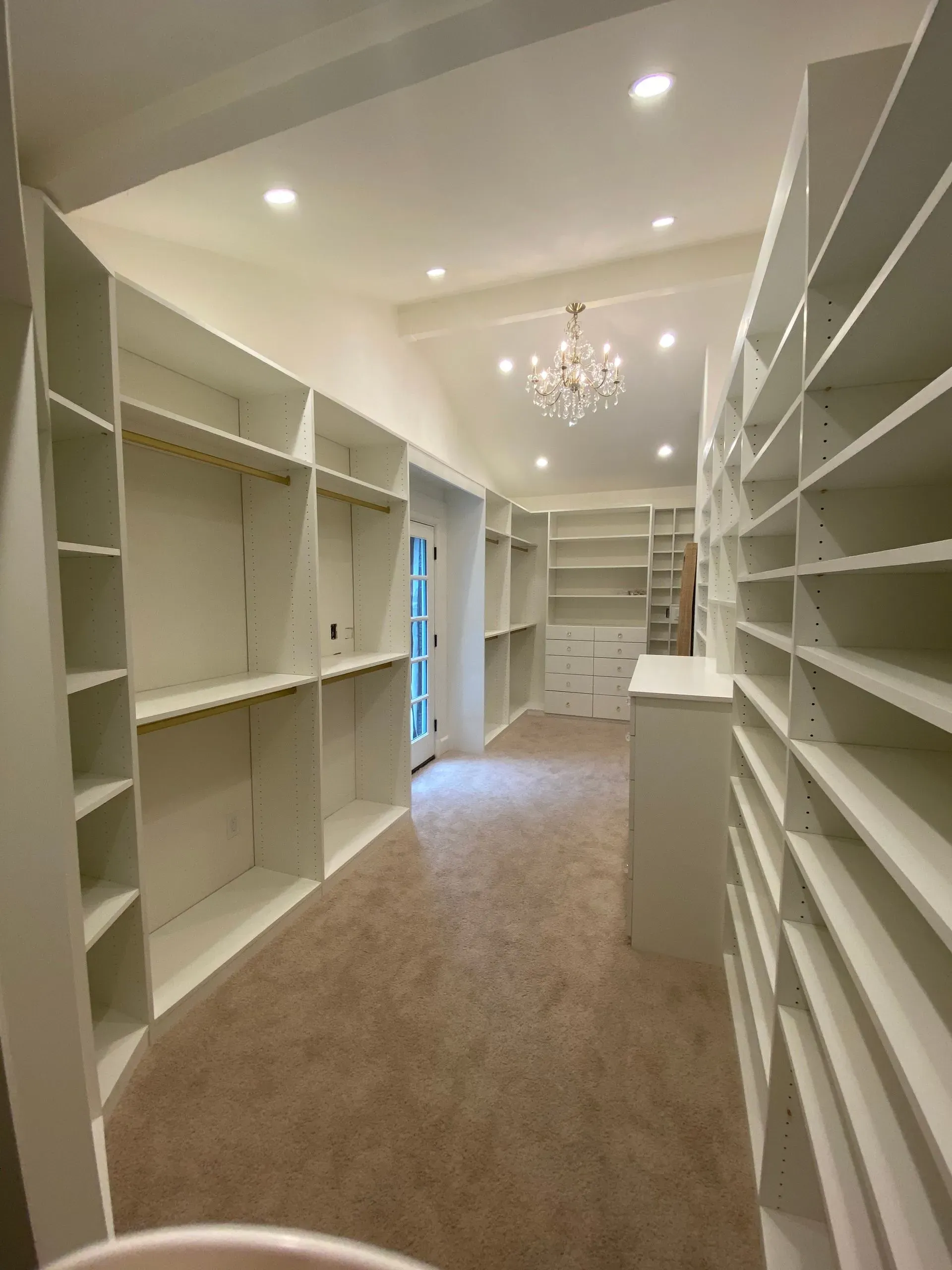 Empty walk-in closet with white shelves, beige carpet, recessed lights, and a chandelier.
