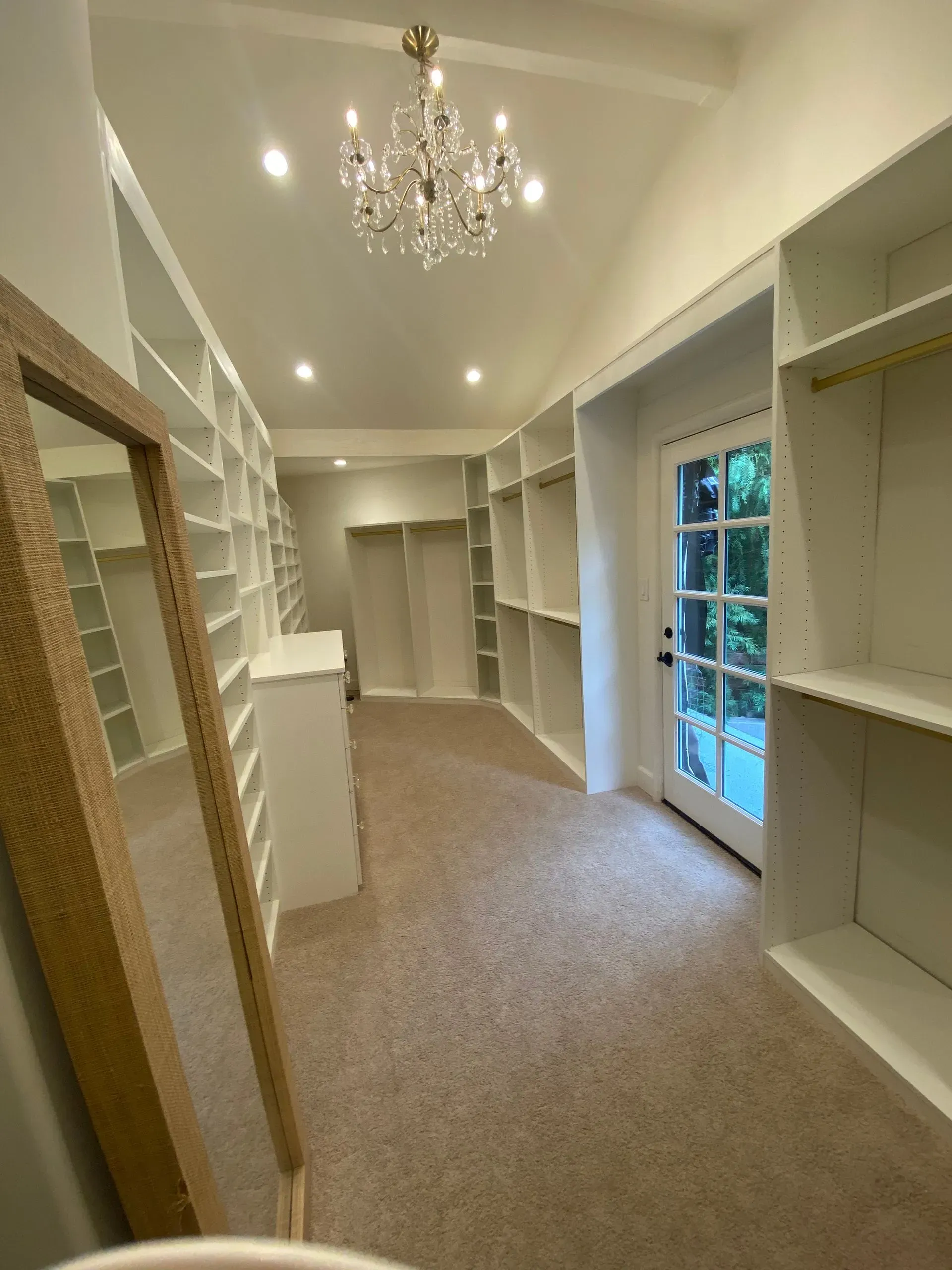 Bright walk-in closet with white built-in shelves, beige carpet, chandelier, and glass doors to the outside.