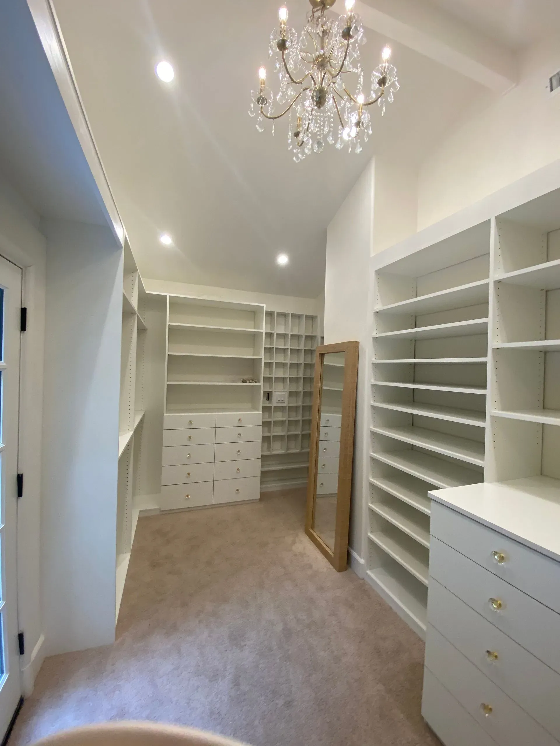 Bright custom walk-in closet with white shelving, drawers, beige carpet, and a chandelier.