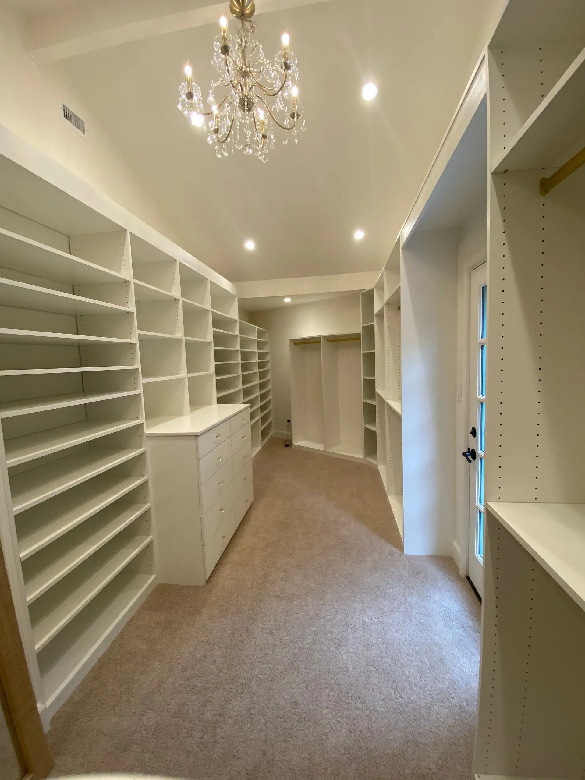 Bright walk-in closet with white shelves, cabinets, carpet, and a chandelier ceiling light.