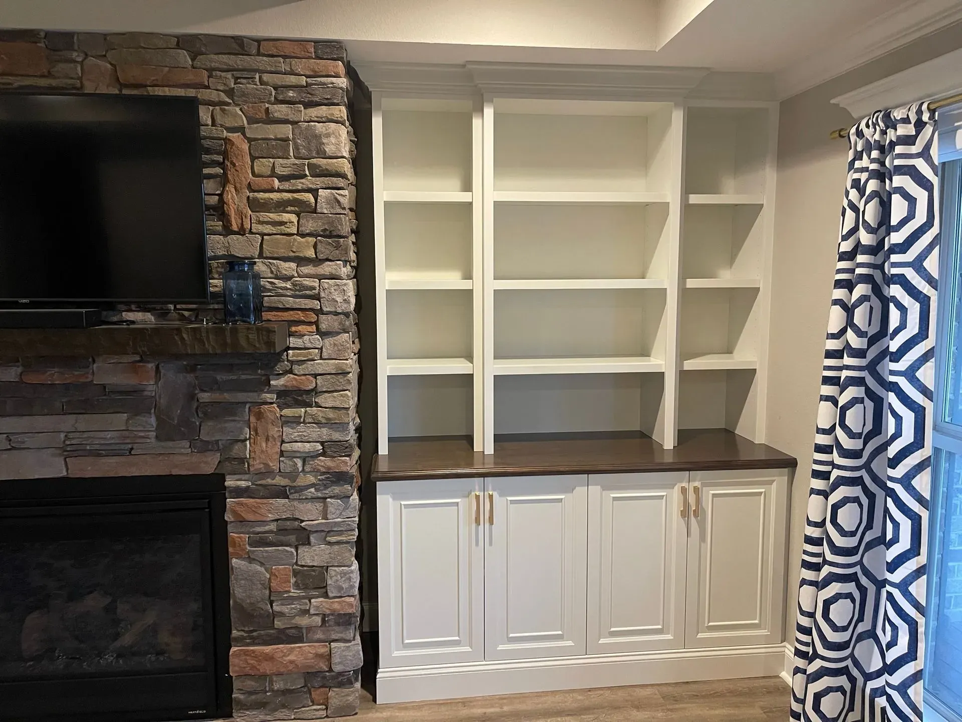 Living room wall with stone fireplace, built-in white shelves, and cabinets beside a patterned curtain