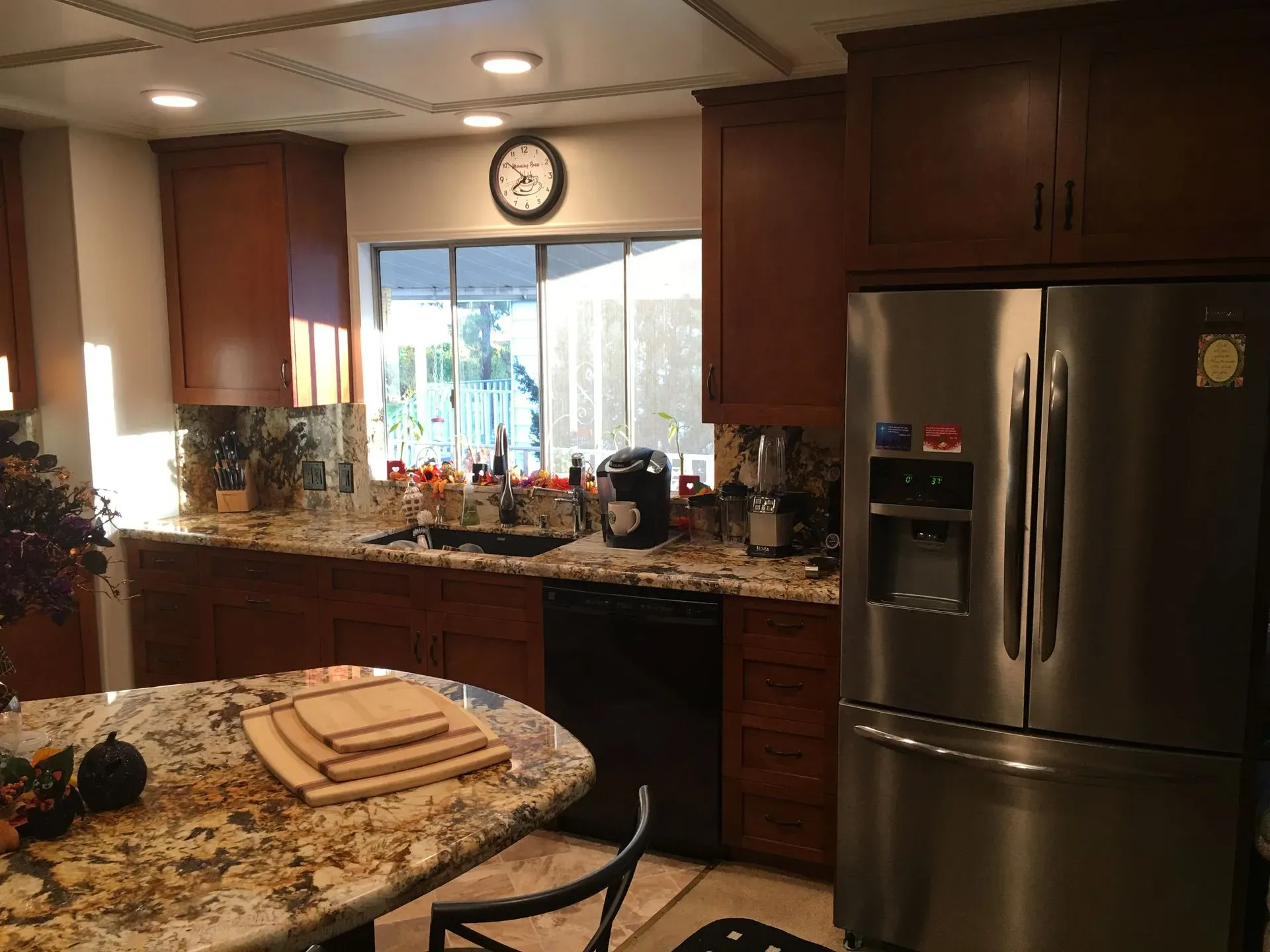 Bright kitchen with wooden cabinets, stainless fridge, granite counters, and a dining table by a window.