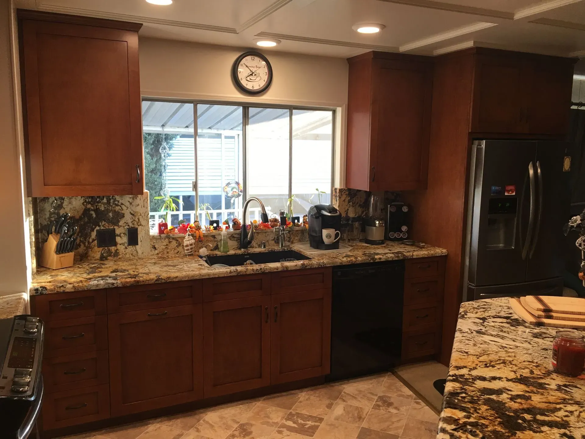 Kitchen with wood cabinets, granite counters, sink under window, clock above, and a black refrigerator.