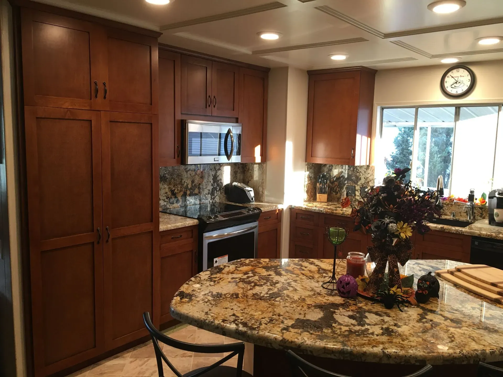 Warm kitchen with wood cabinets, granite island, stainless stove, and a sunlit window with plants and clock.