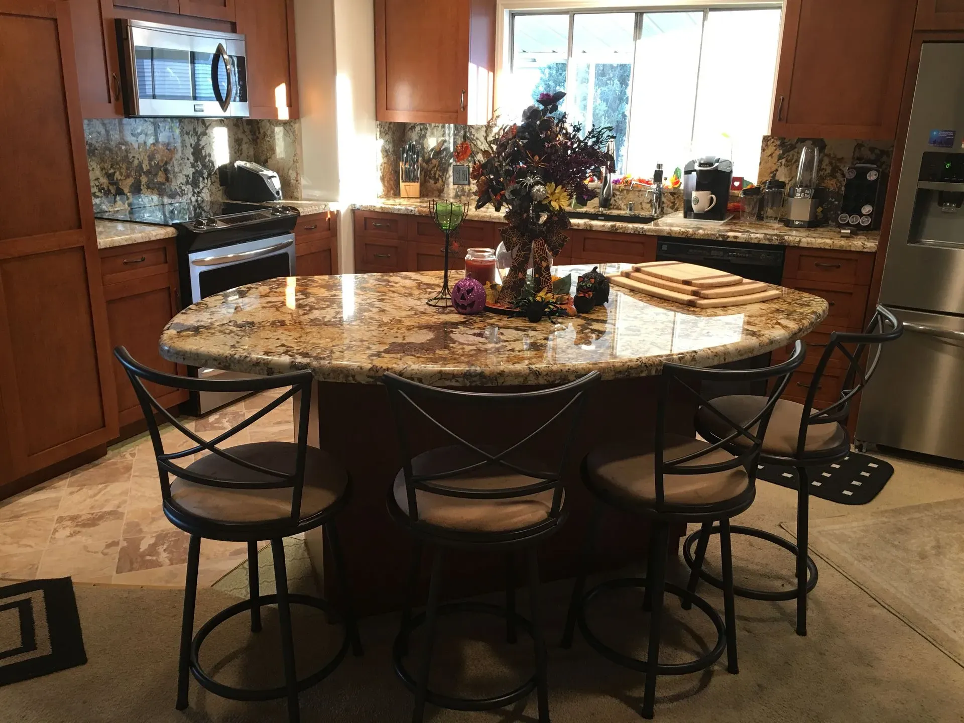 Kitchen with a granite island, four stools, and a floral centerpiece, lit by sunlight through a window.
