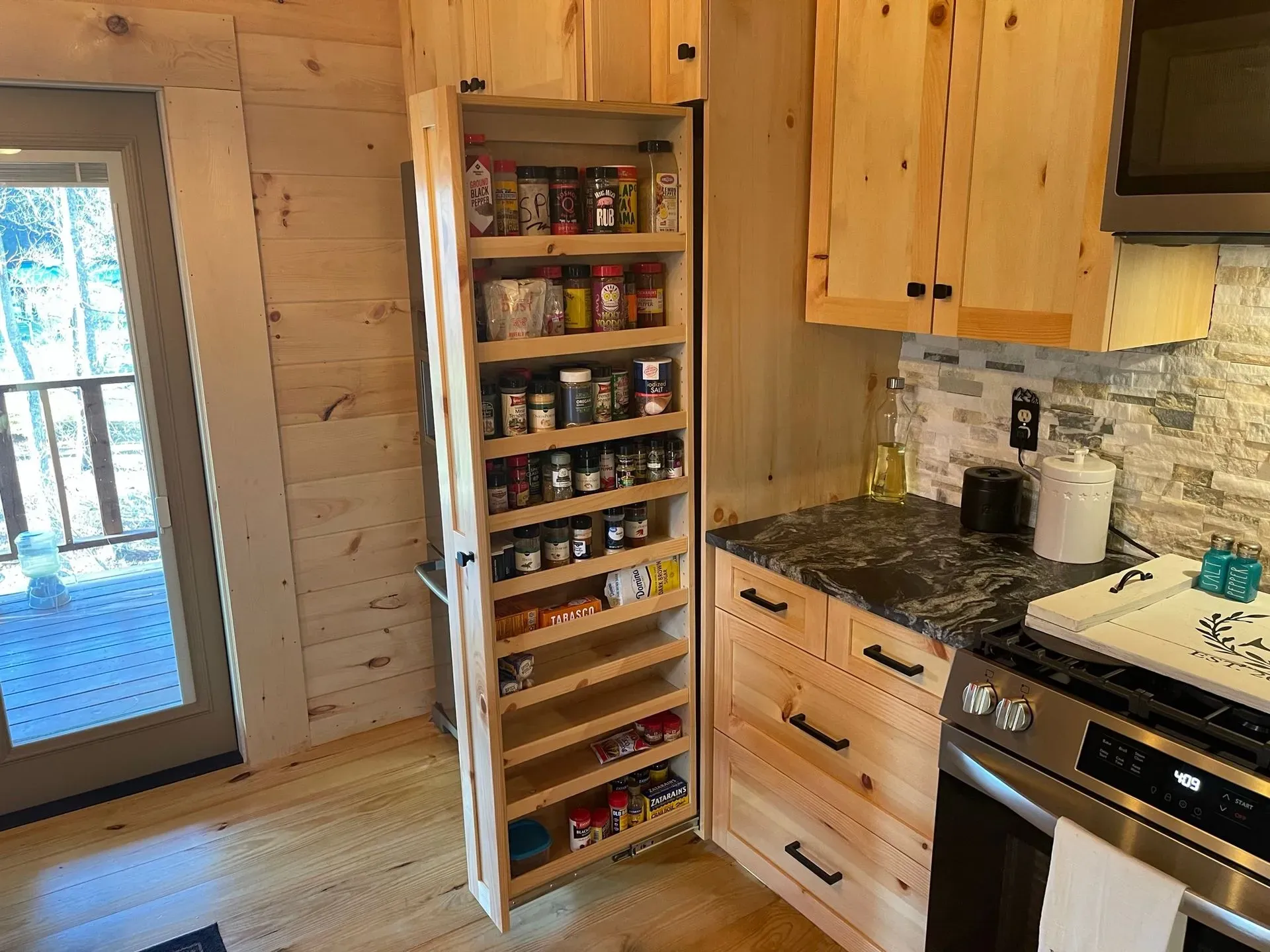 Rustic kitchen corner with open spice shelf, wooden cabinets, granite counter, and glass door to deck.