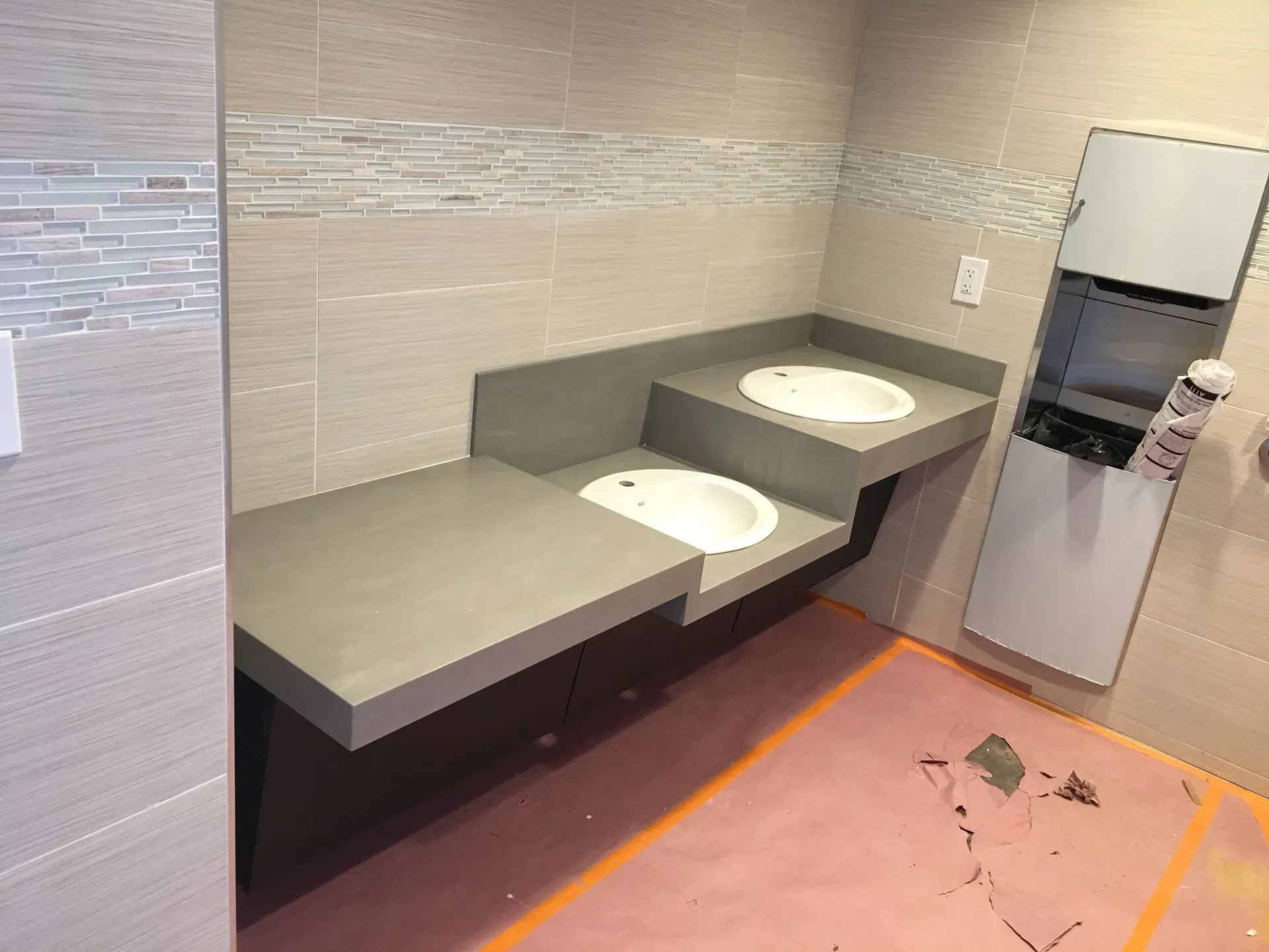 Public restroom with two gray sinks, beige tiled walls, and a stainless hand dryer.