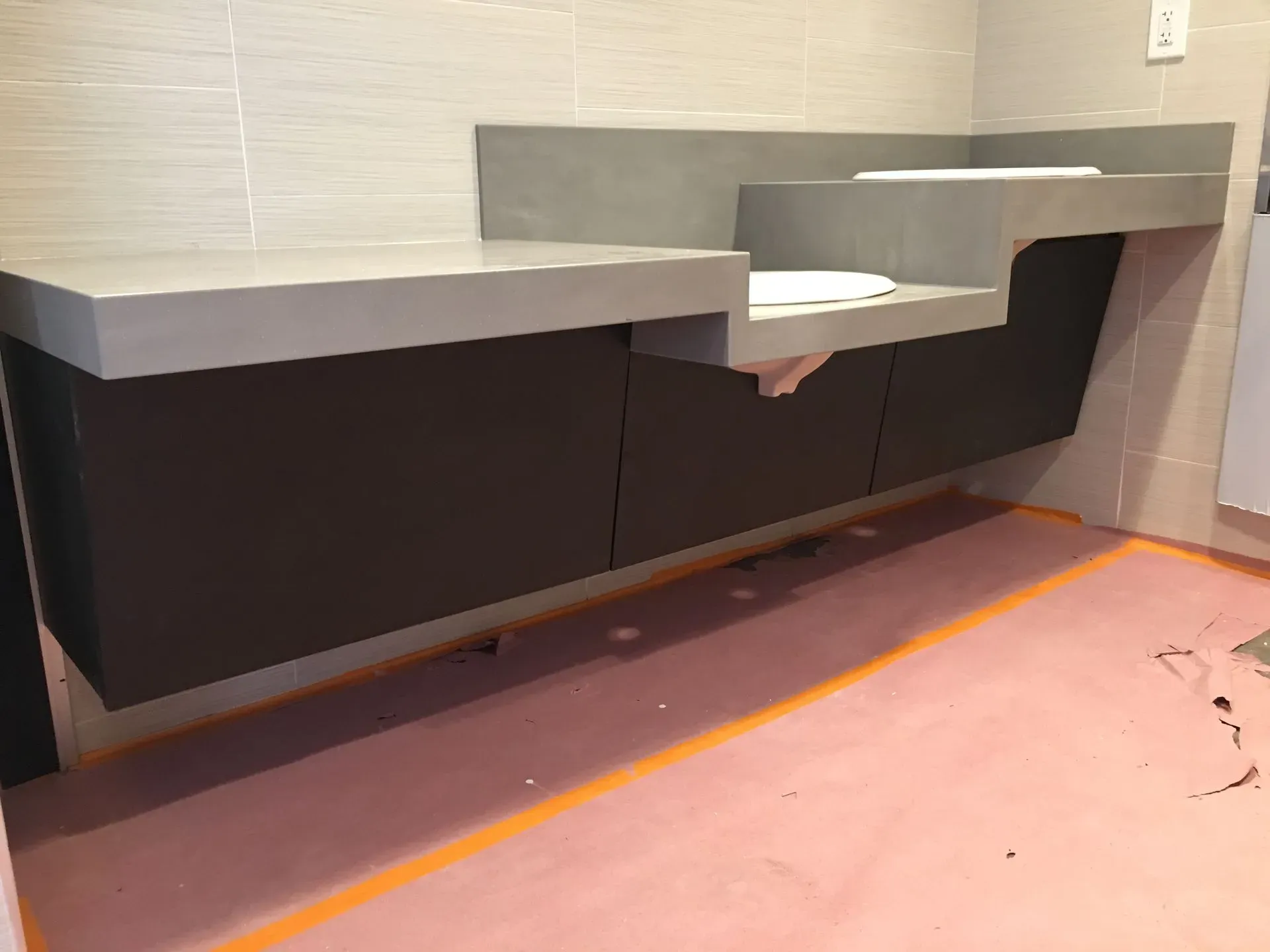 Modern restroom with a long dark vanity, gray countertop, and orange floor protection tape.
