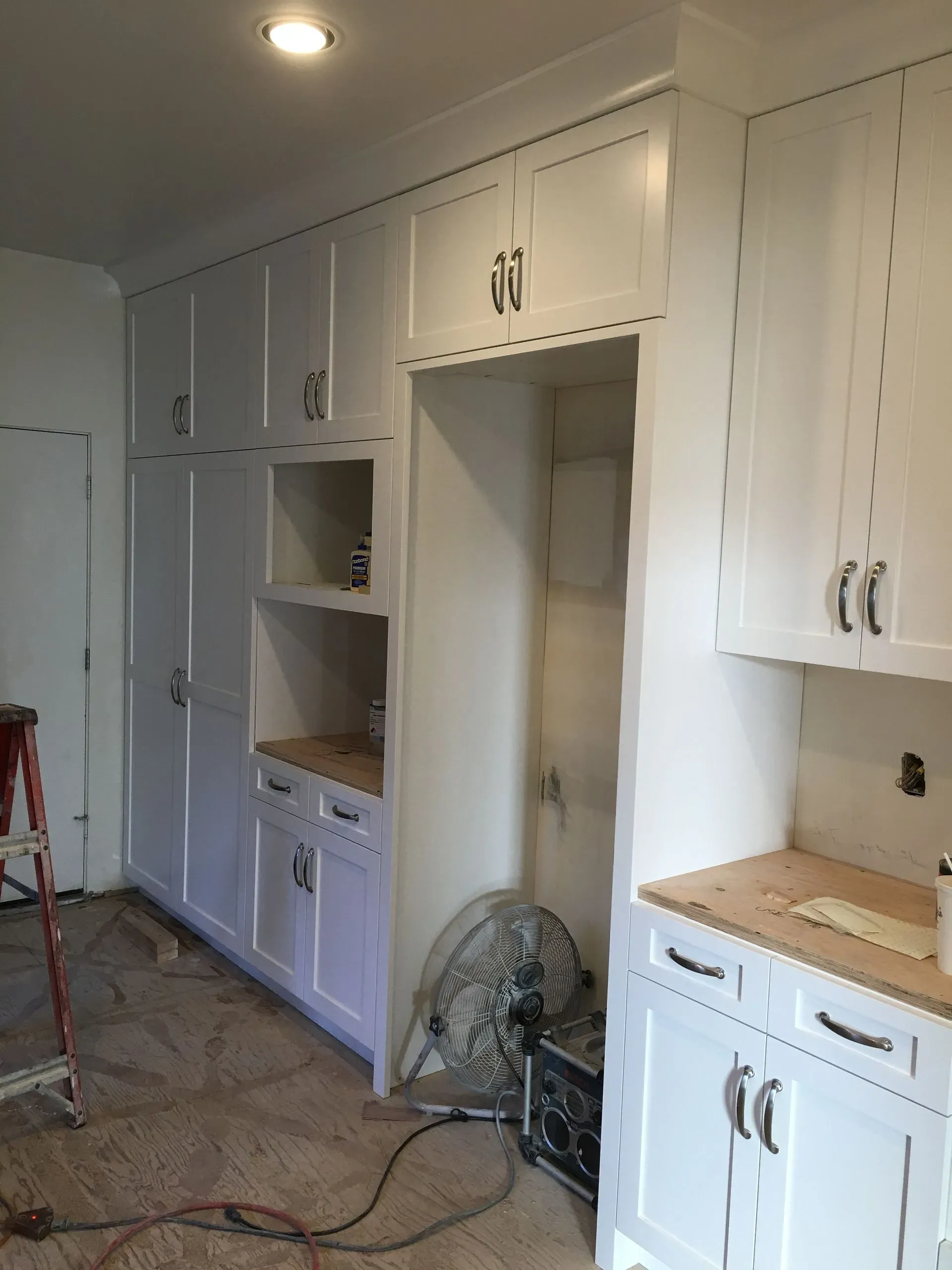 Renovated kitchen with white cabinets, built-in shelves, and a floor fan; tools and cords on the floor.