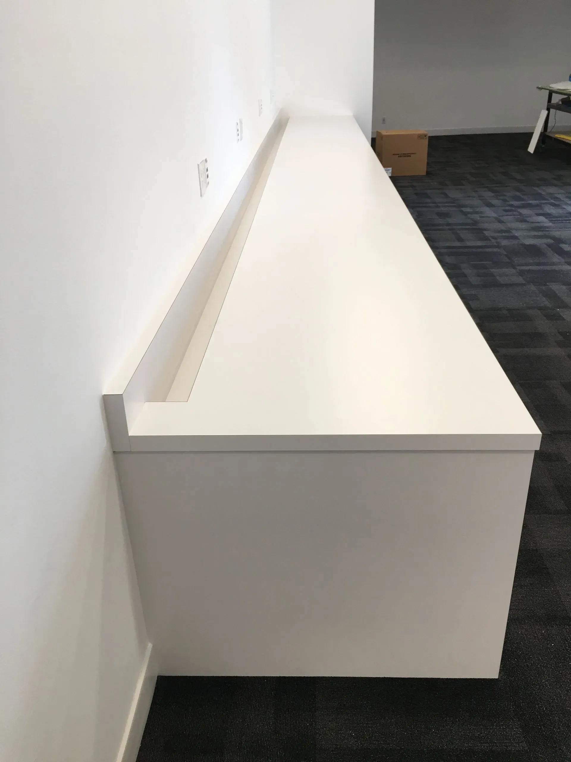Long white wall-mounted shelf or console in an office hallway against a white wall and carpeted floor.