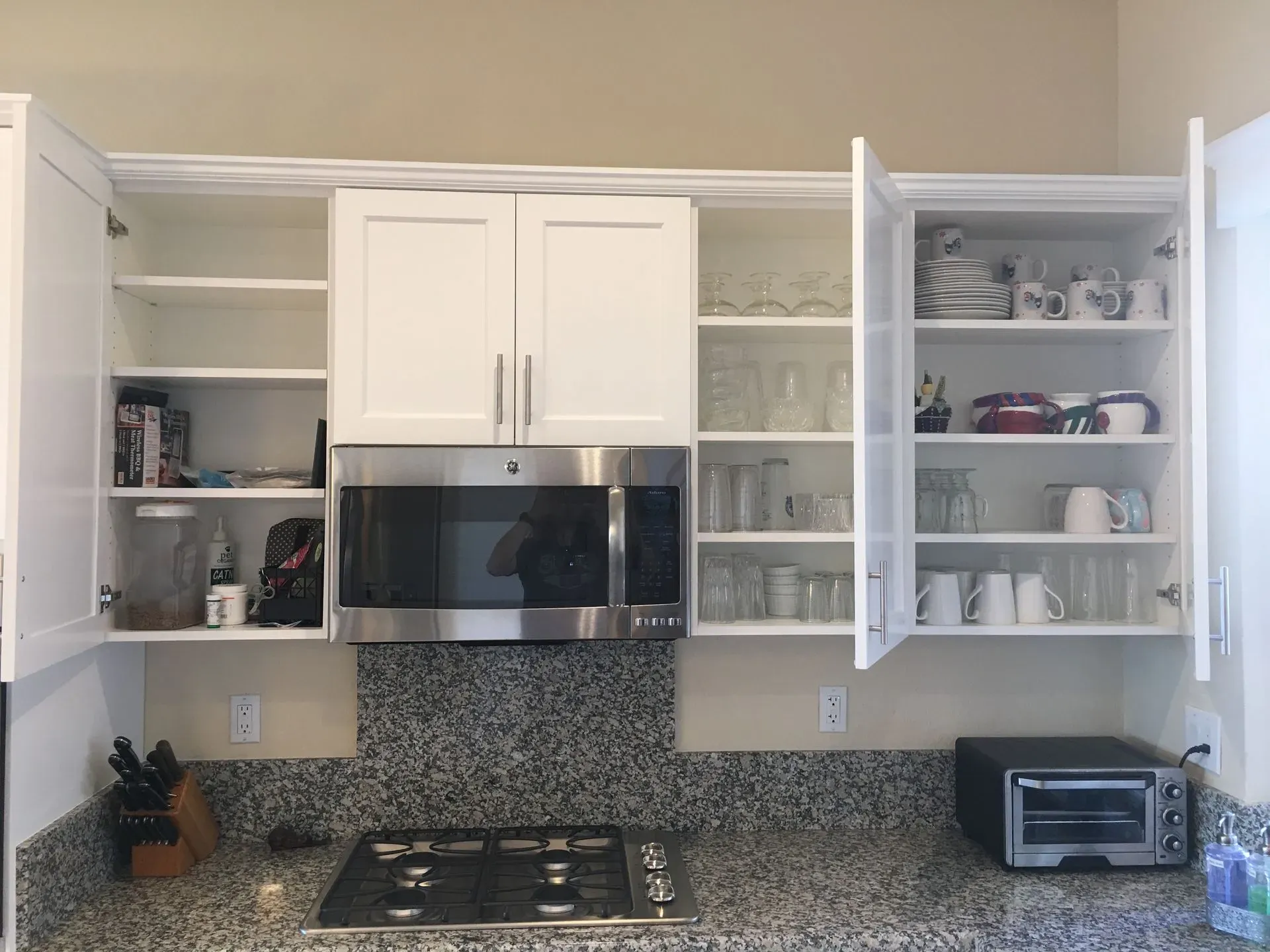 White kitchen with open cabinets, microwave, coffee mugs, and a gas stove with speckled backsplash.
