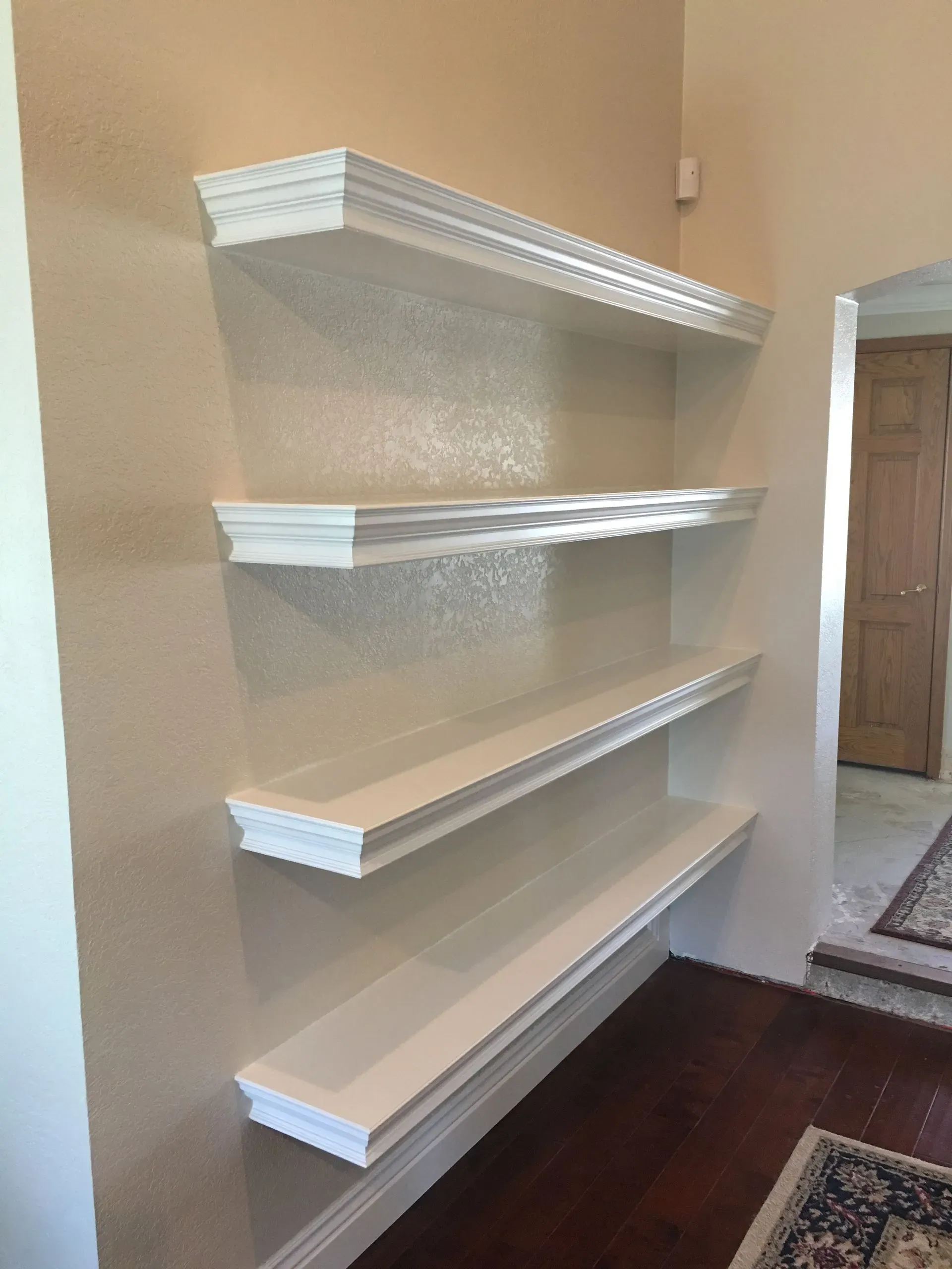 Four white floating shelves mounted on a beige hallway wall above dark wood flooring.