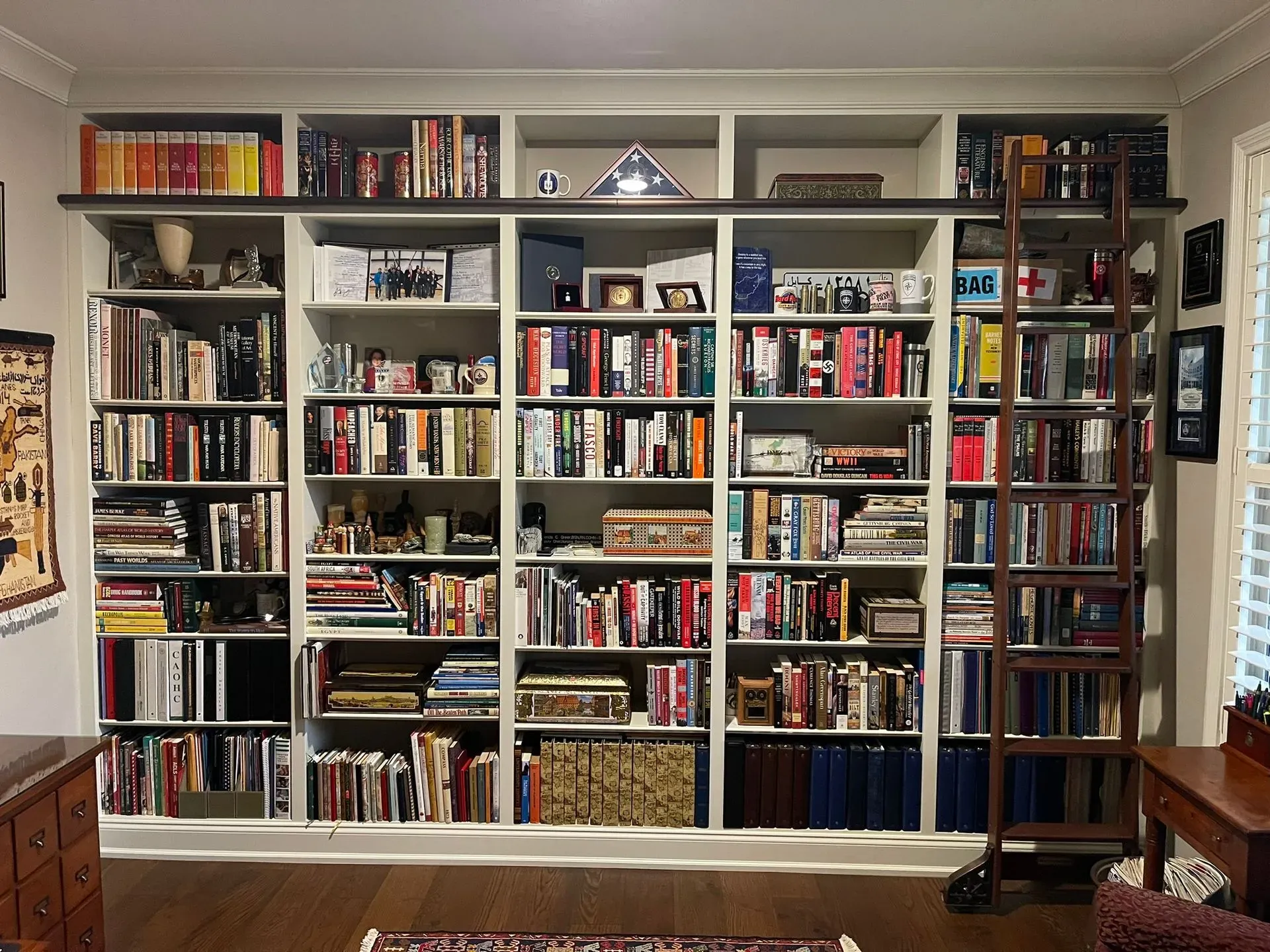 Wall of bookshelves filled with books and decor in a cozy living room, with hardwood floors and a ladder on the right.
