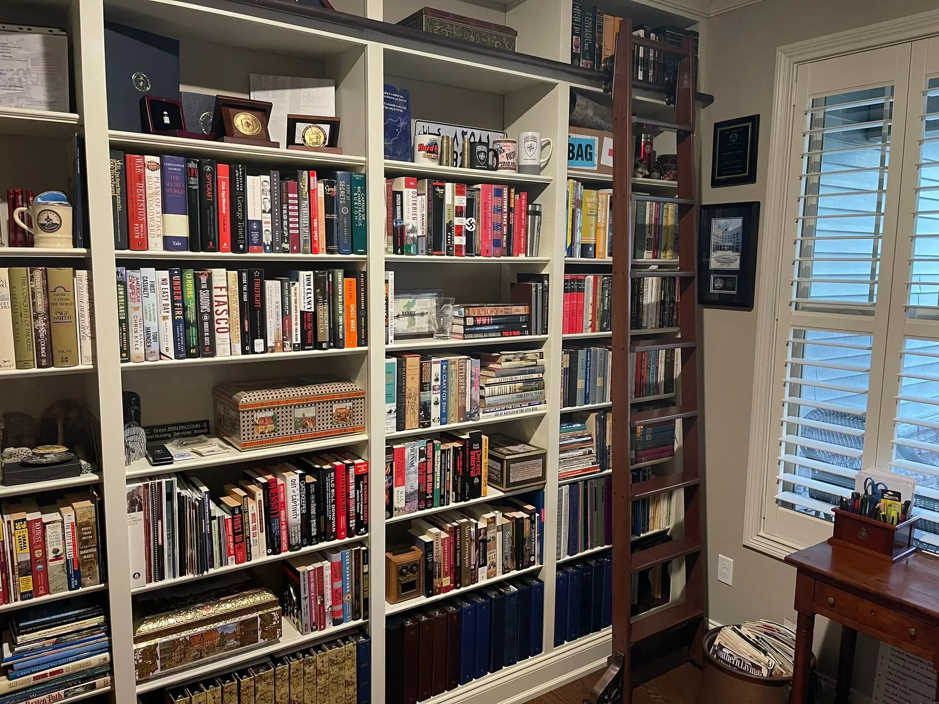 A home office bookshelf filled with books, records, and framed photos beside a window with shutters.